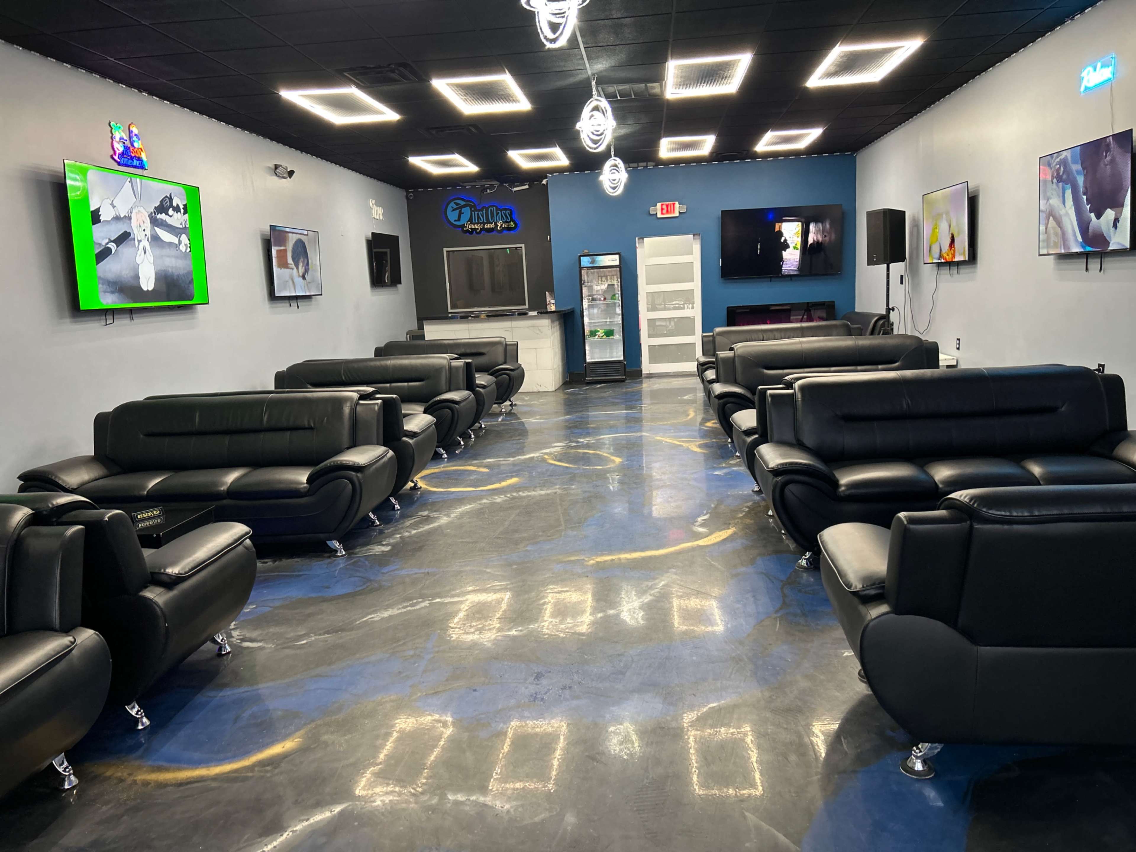 A lounge area with black leather couches arranged around a central space, illuminated by bright ceiling lights and featuring several mounted televisions on the walls.