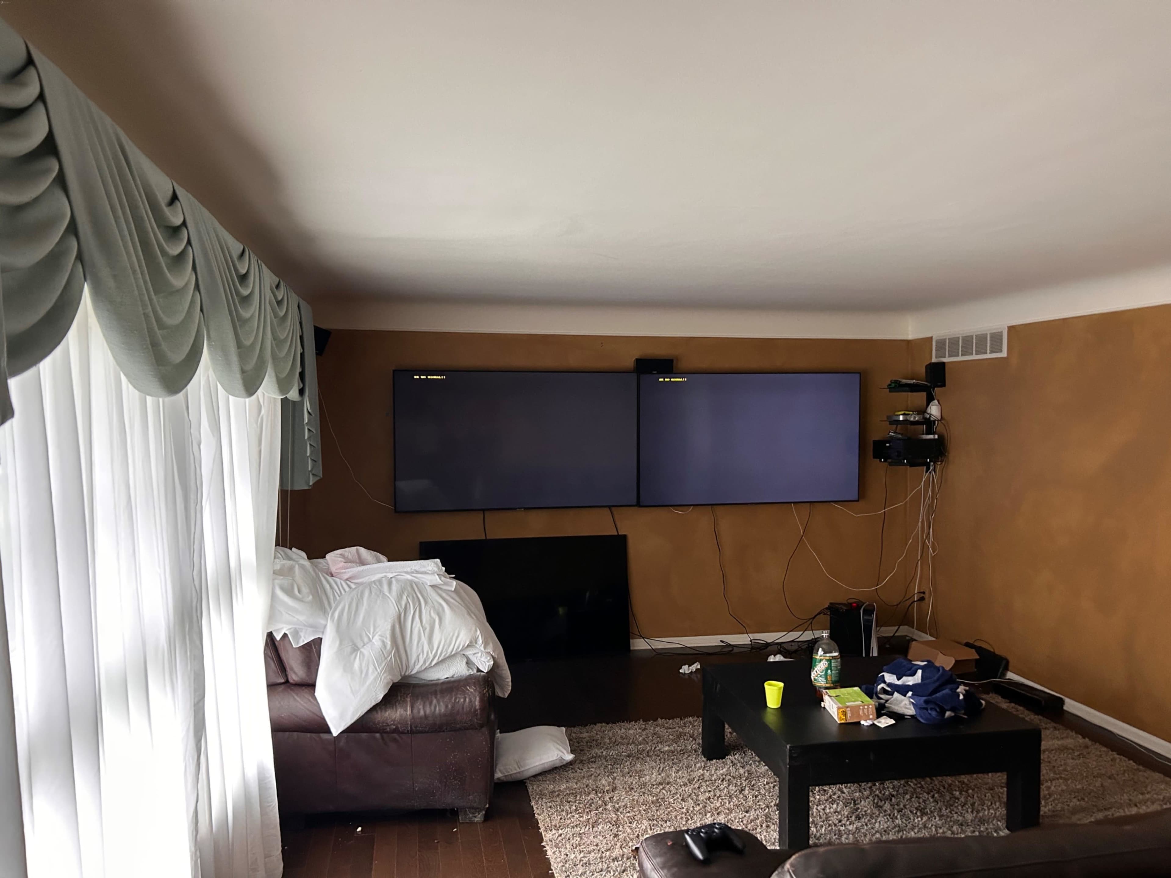 A living room features two mounted flat-screen TVs on a wall, a brown couch with a blanket and clothes, and a rug on the wooden floor.