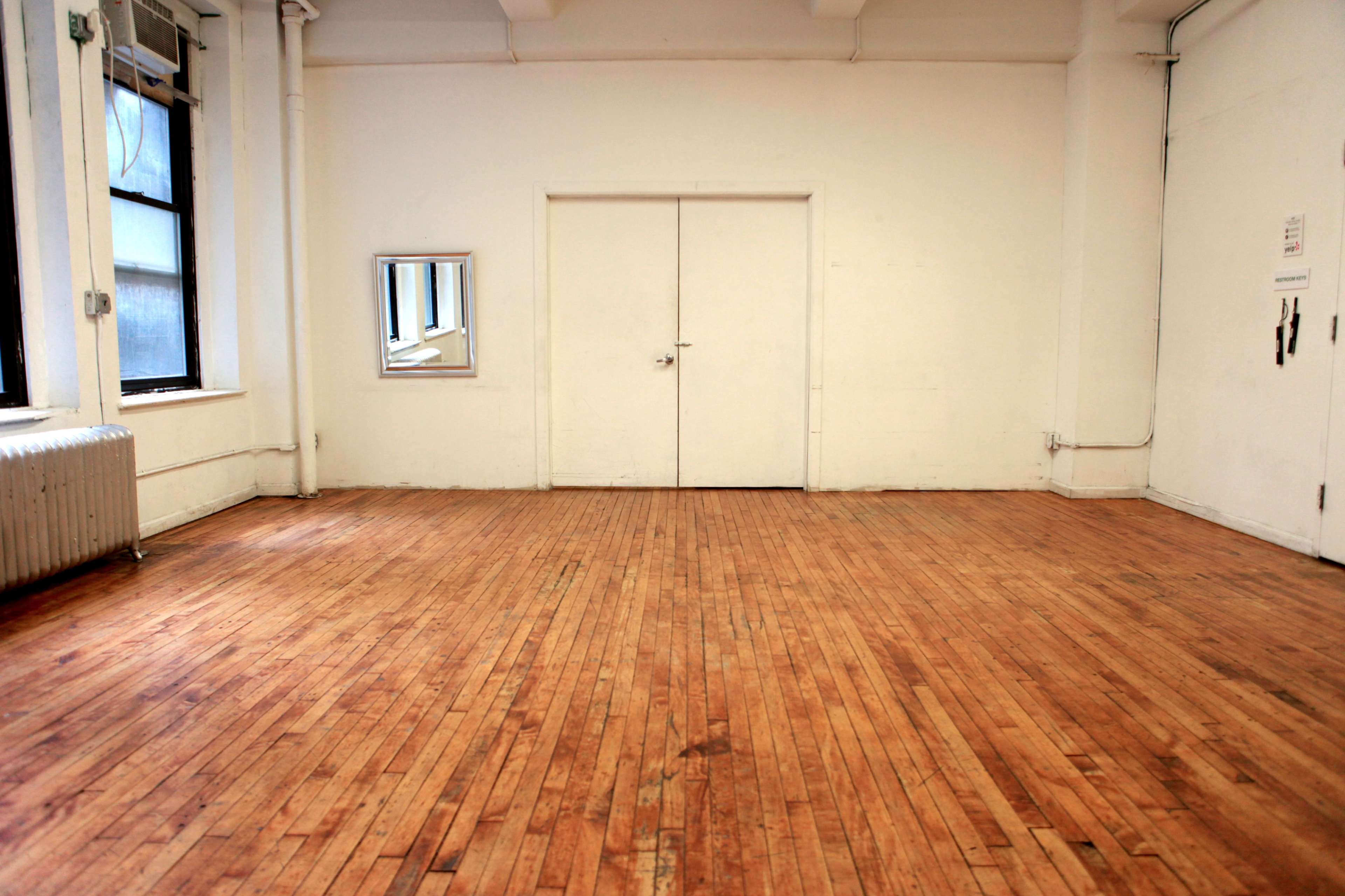 A spacious, empty room with wooden floors, white walls, a mirror, and a closed door.