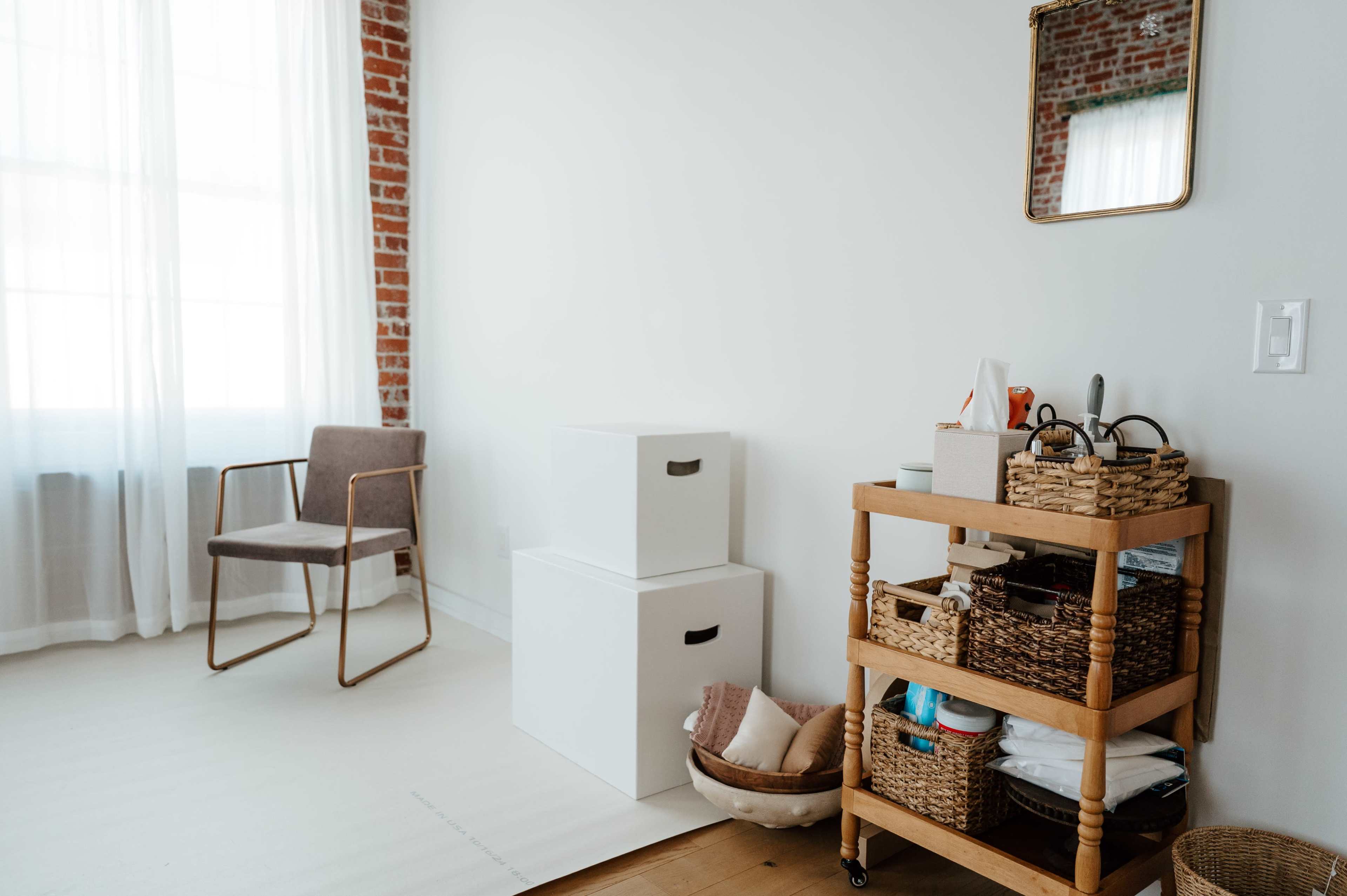 The image shows a bright room with a window, featuring a chair, stacked white boxes, and a wooden cart with woven baskets.
