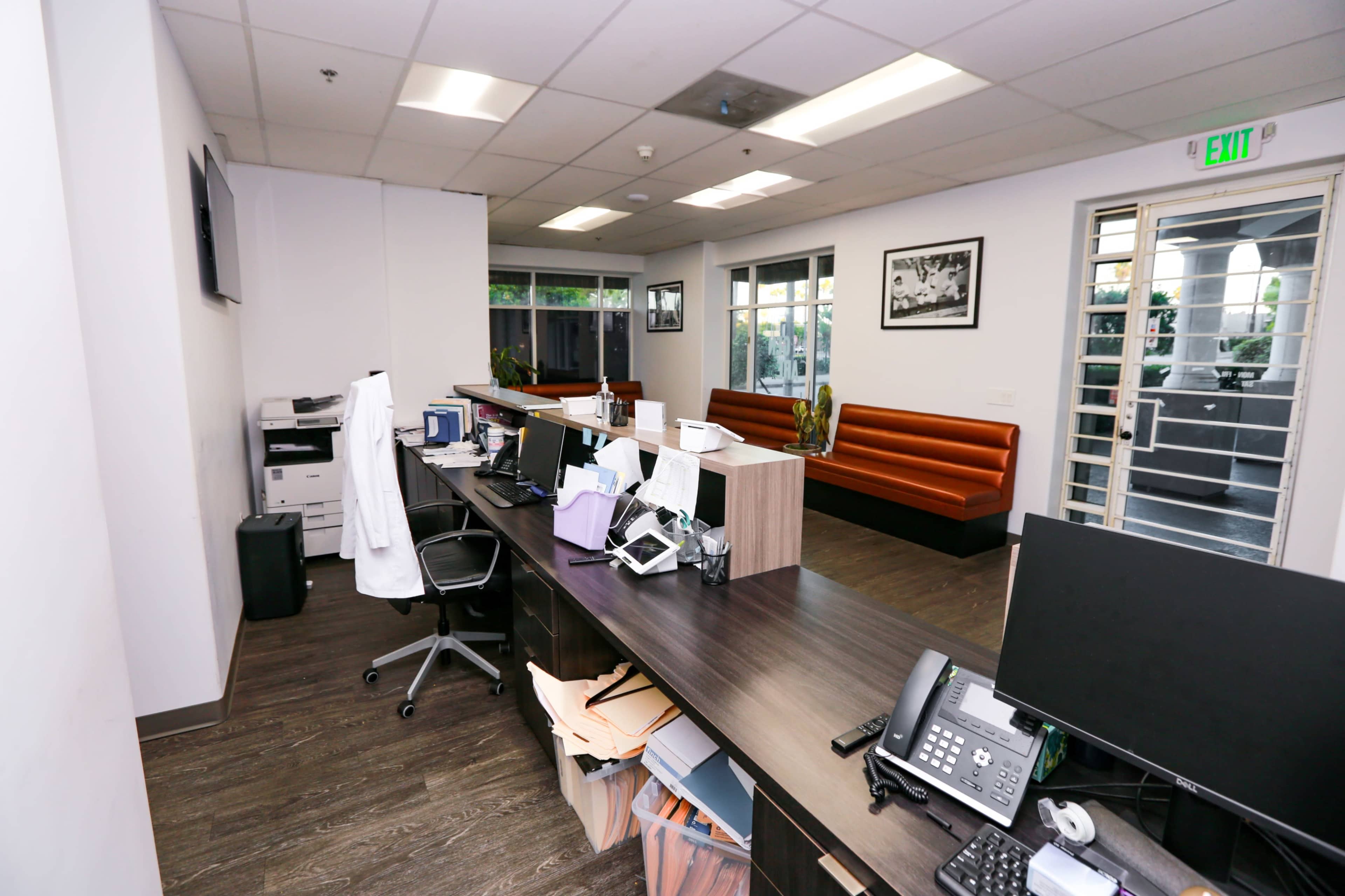 The image shows a reception area of an office with a desk, printer, and seating along with a large window, allowing natural light to enter.