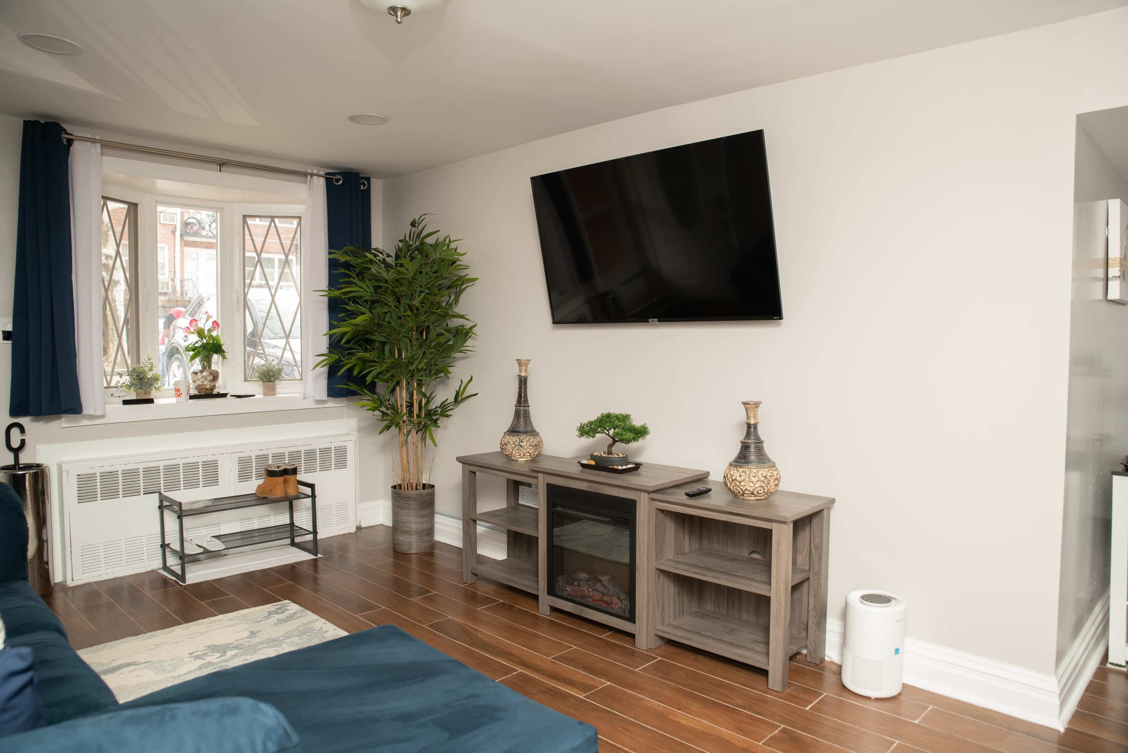 A living room with a mounted television on the wall, a couch, decorative plants, and a coffee table underneath a window.