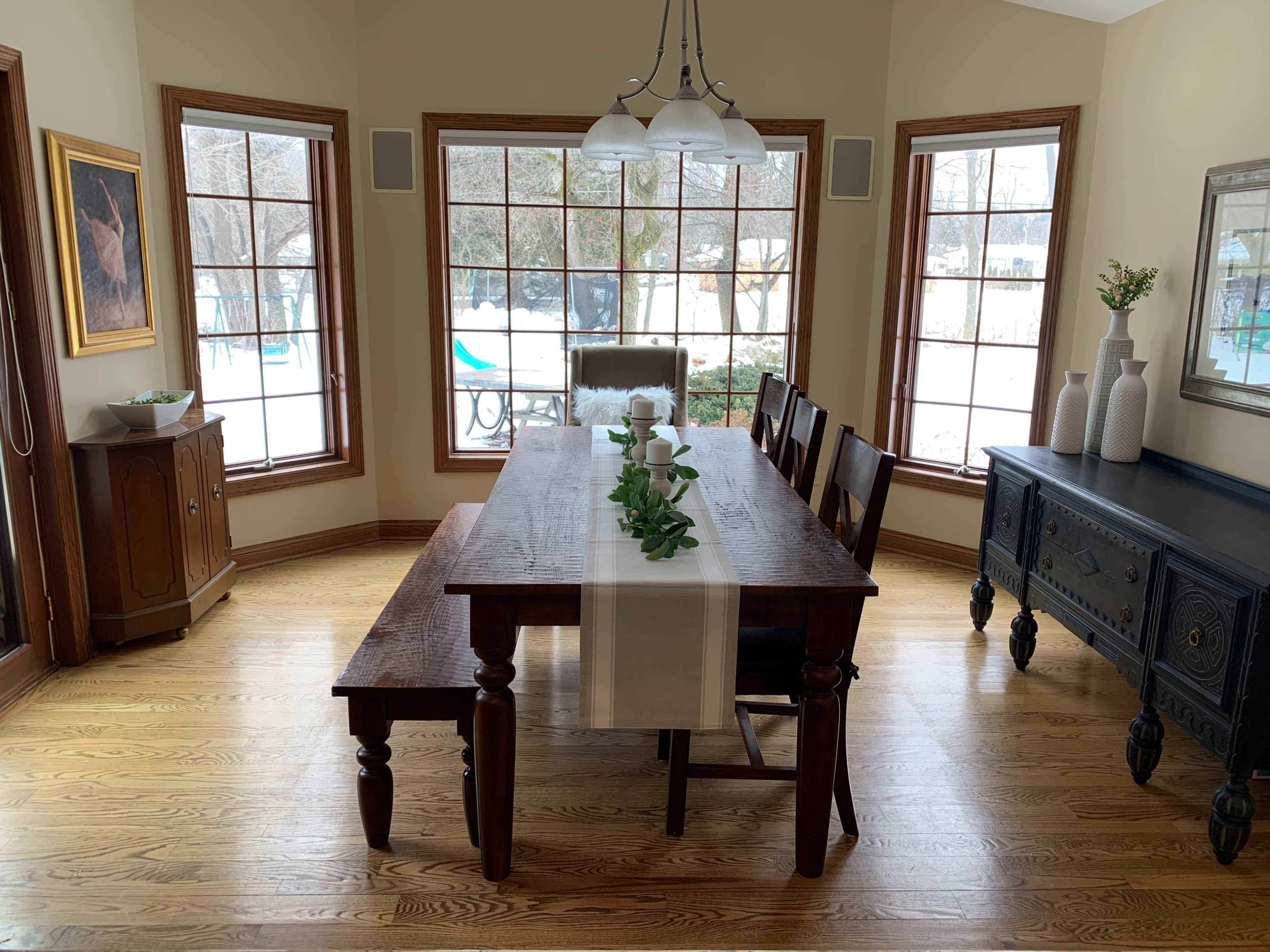 A wooden dining table with a runner and greenery sits in a well-lit room featuring large windows and a view of an outdoor space.