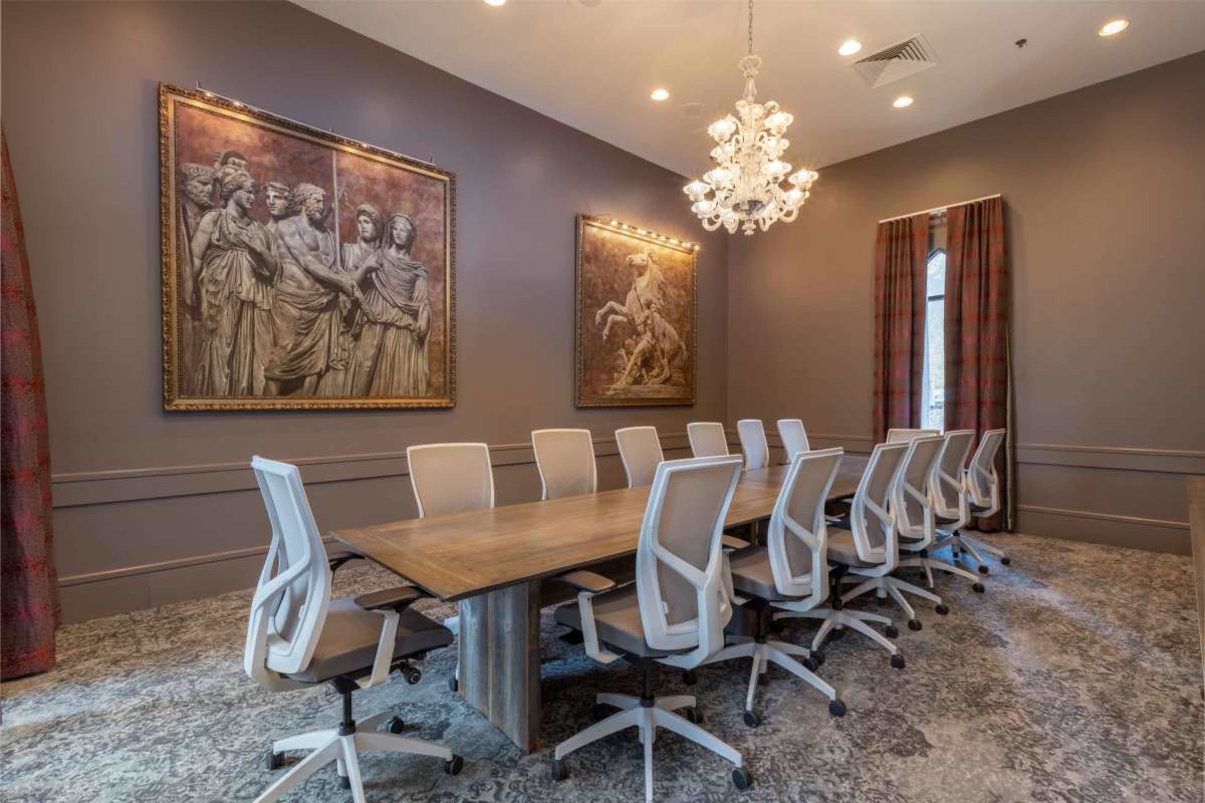 A conference room features a long wooden table surrounded by white office chairs and adorned with art pieces and a chandelier.