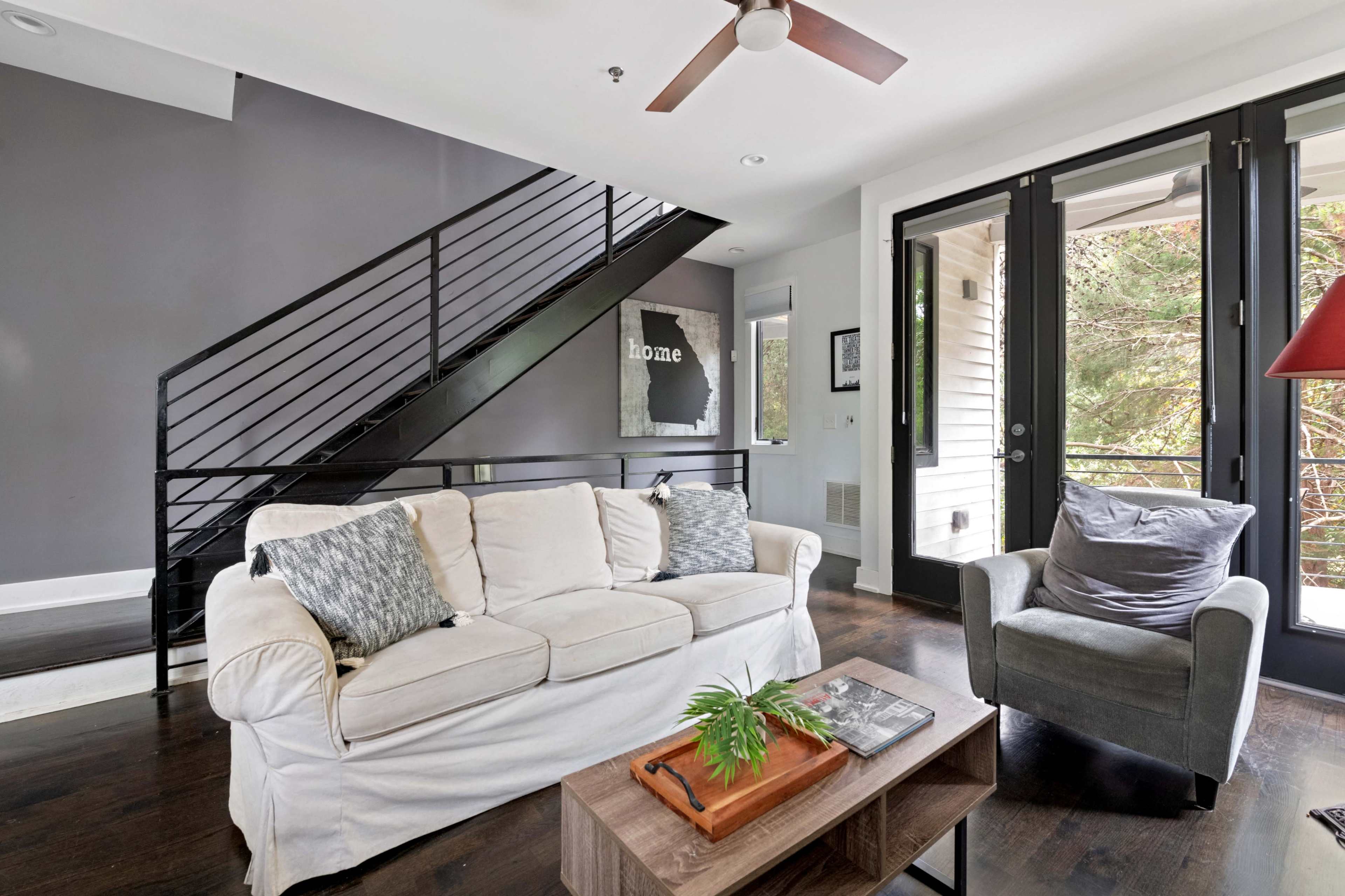 A modern living room features a white sofa, a gray armchair, and a wooden coffee table, with a staircase and large windows providing natural light.