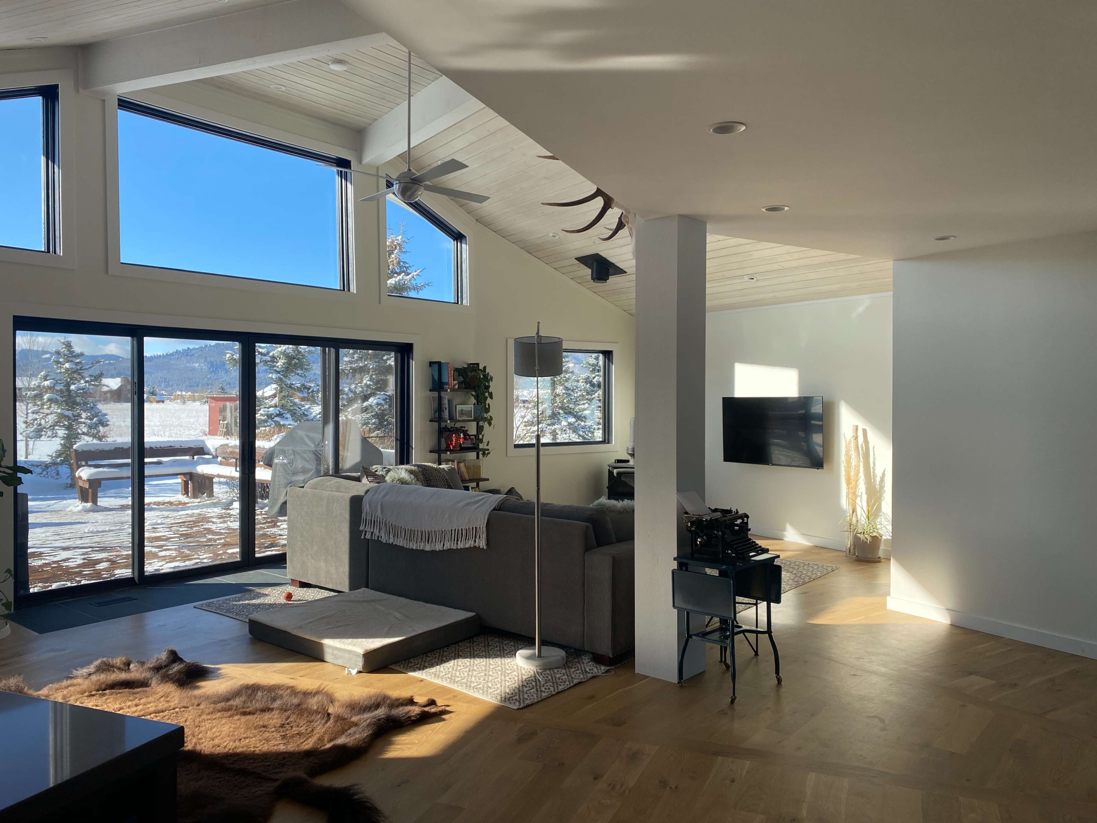 A modern living room with large windows overlooking a snowy landscape, furnished with a gray sofa and a television mounted on the wall.