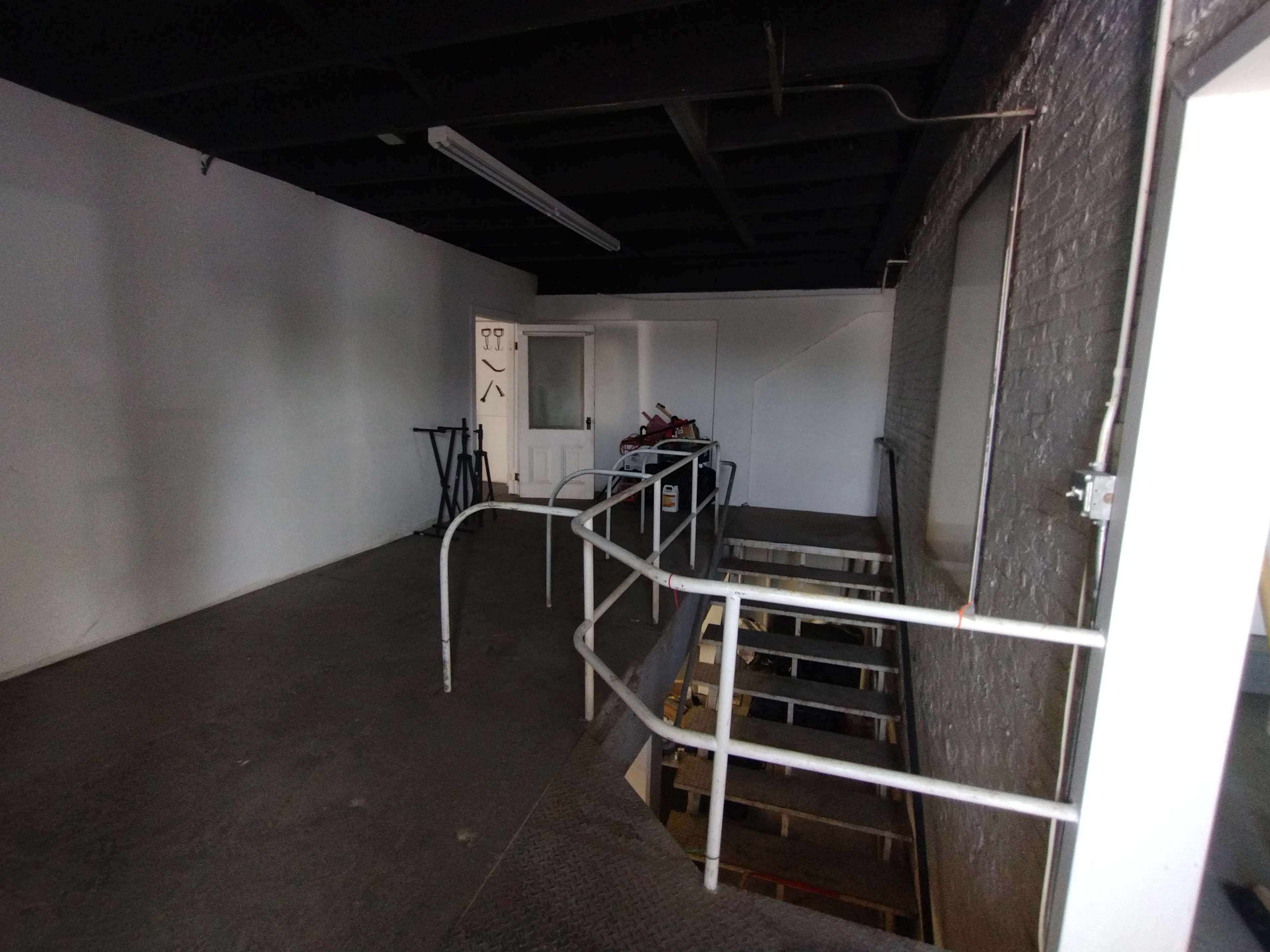A dimly lit staircase leading down from a white-walled room, with a railing on the left and a small entryway at the top.