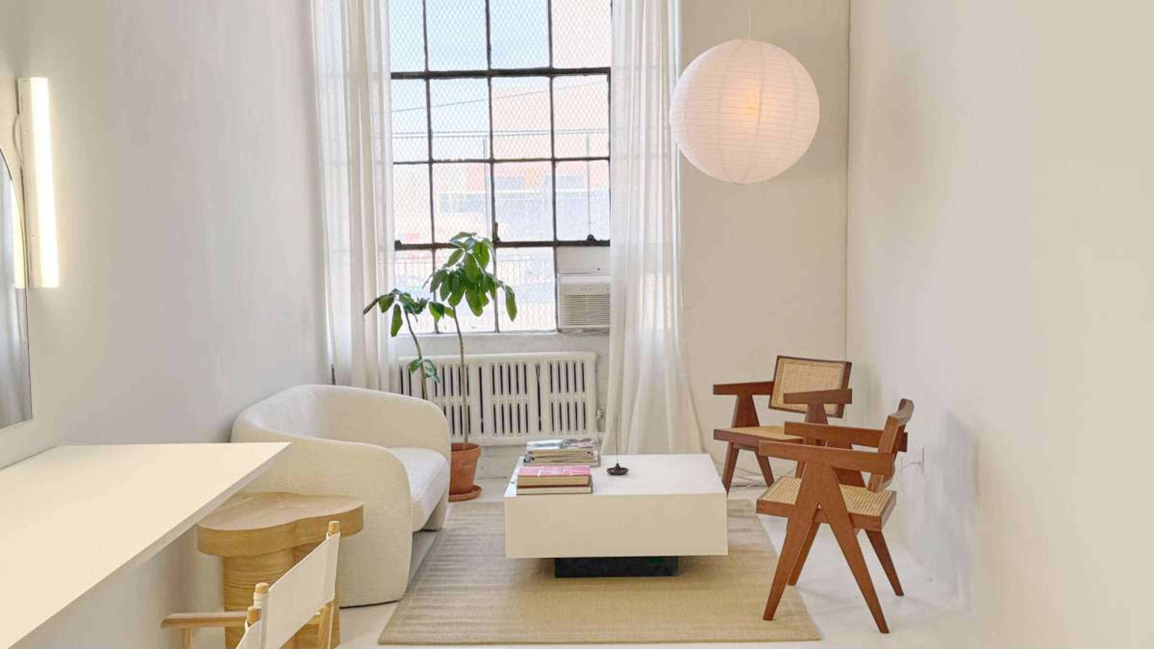The image shows a minimalist living space with light-colored walls, a large window, a potted plant, and a few modern furniture pieces, including a white chair, a wooden table, and two wooden chairs.