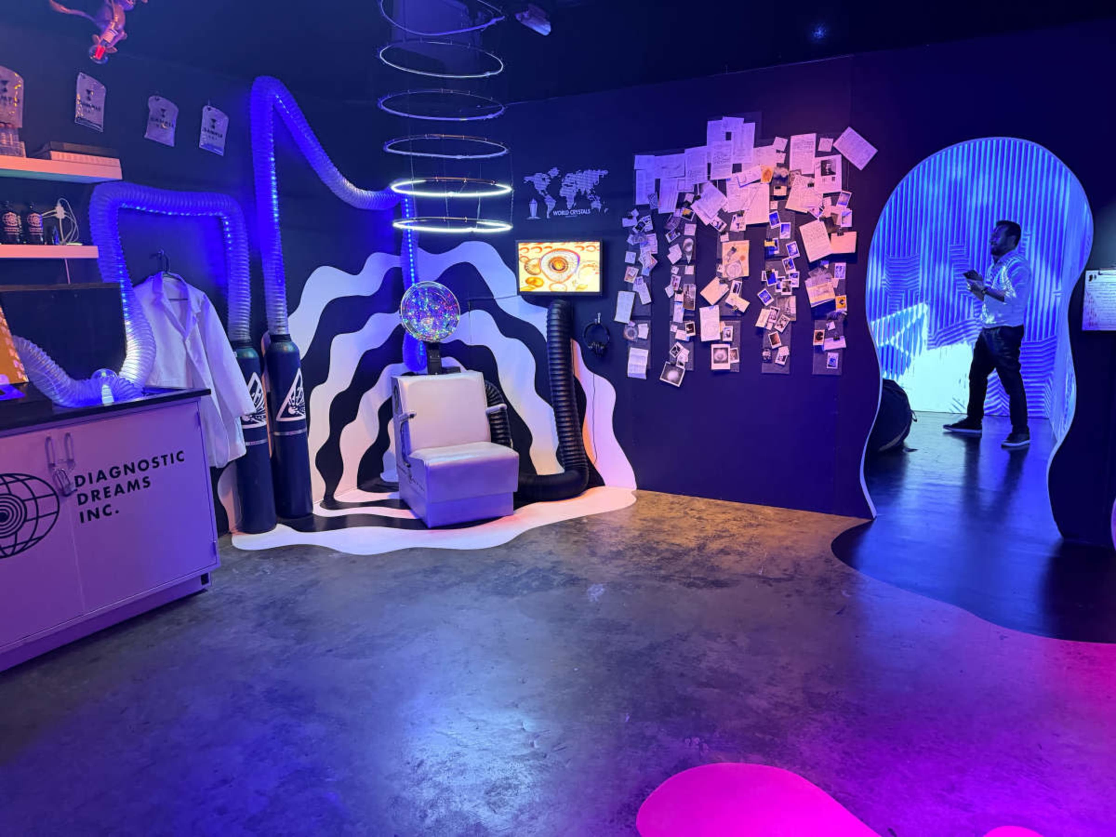 The image shows a modern, colorful exhibition space featuring abstract designs, a white chair, a display screen, and various wall-mounted elements, including photographs and papers.