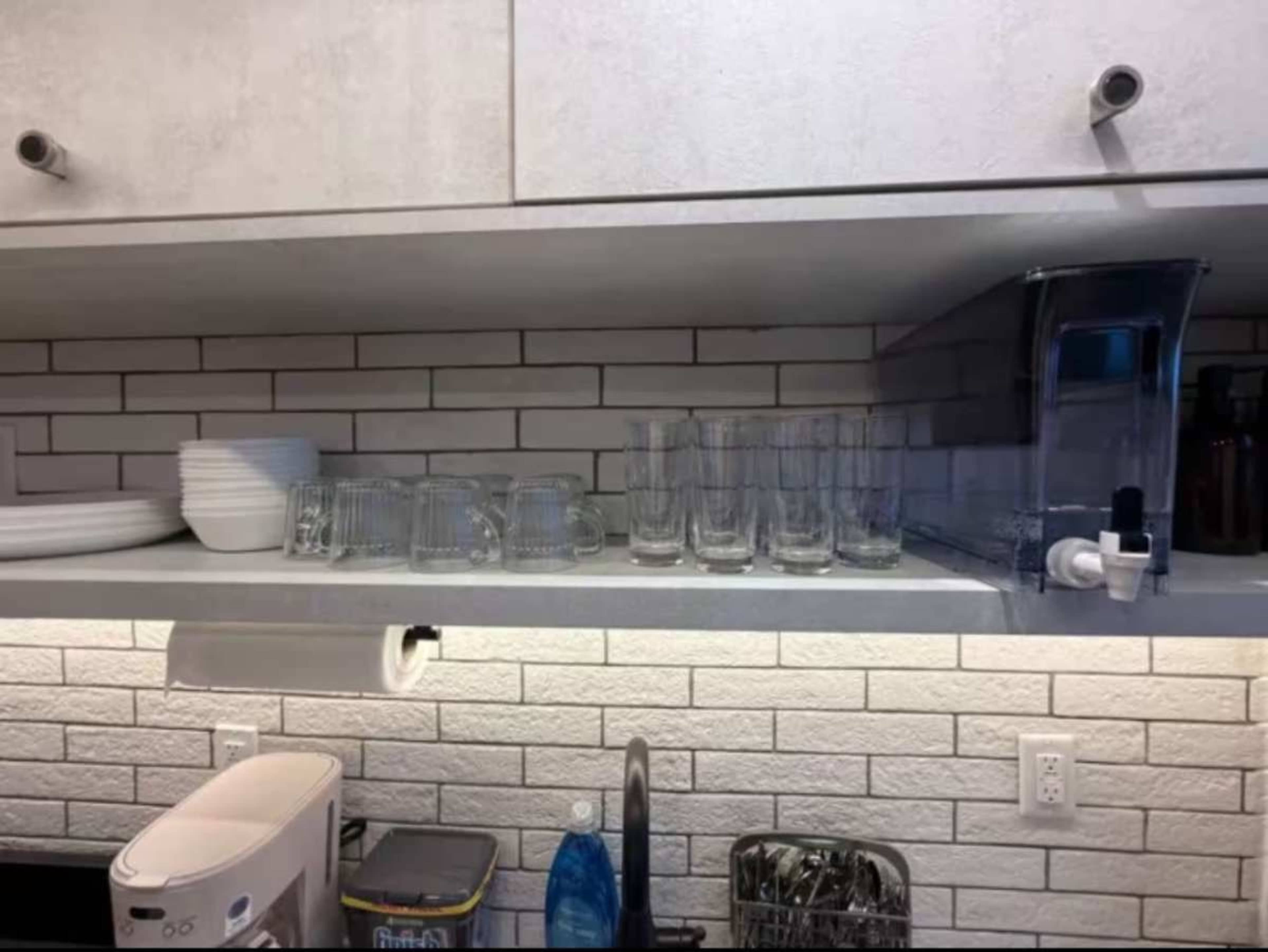 The image shows a kitchen shelf with stacked white plates, several clear glass cups, and a water dispenser, all arranged in a clean, organized manner.