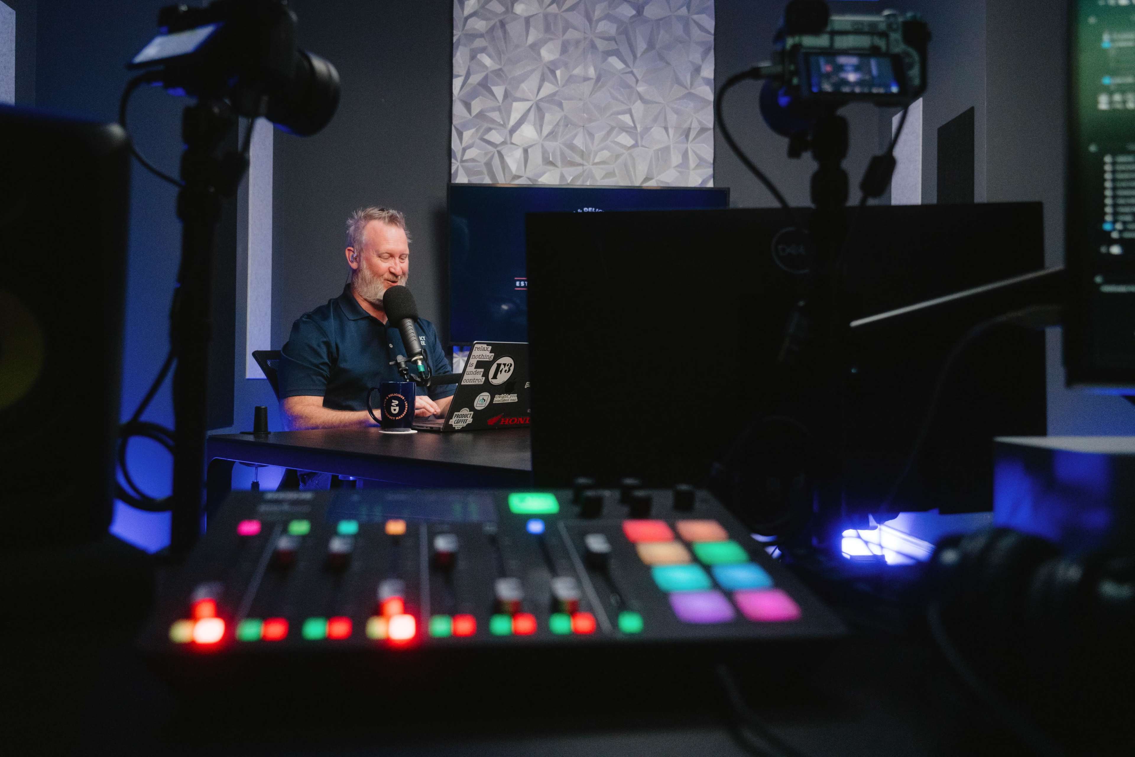 A person is sitting at a desk with a microphone and multiple cameras set up, surrounded by screens and audio equipment.