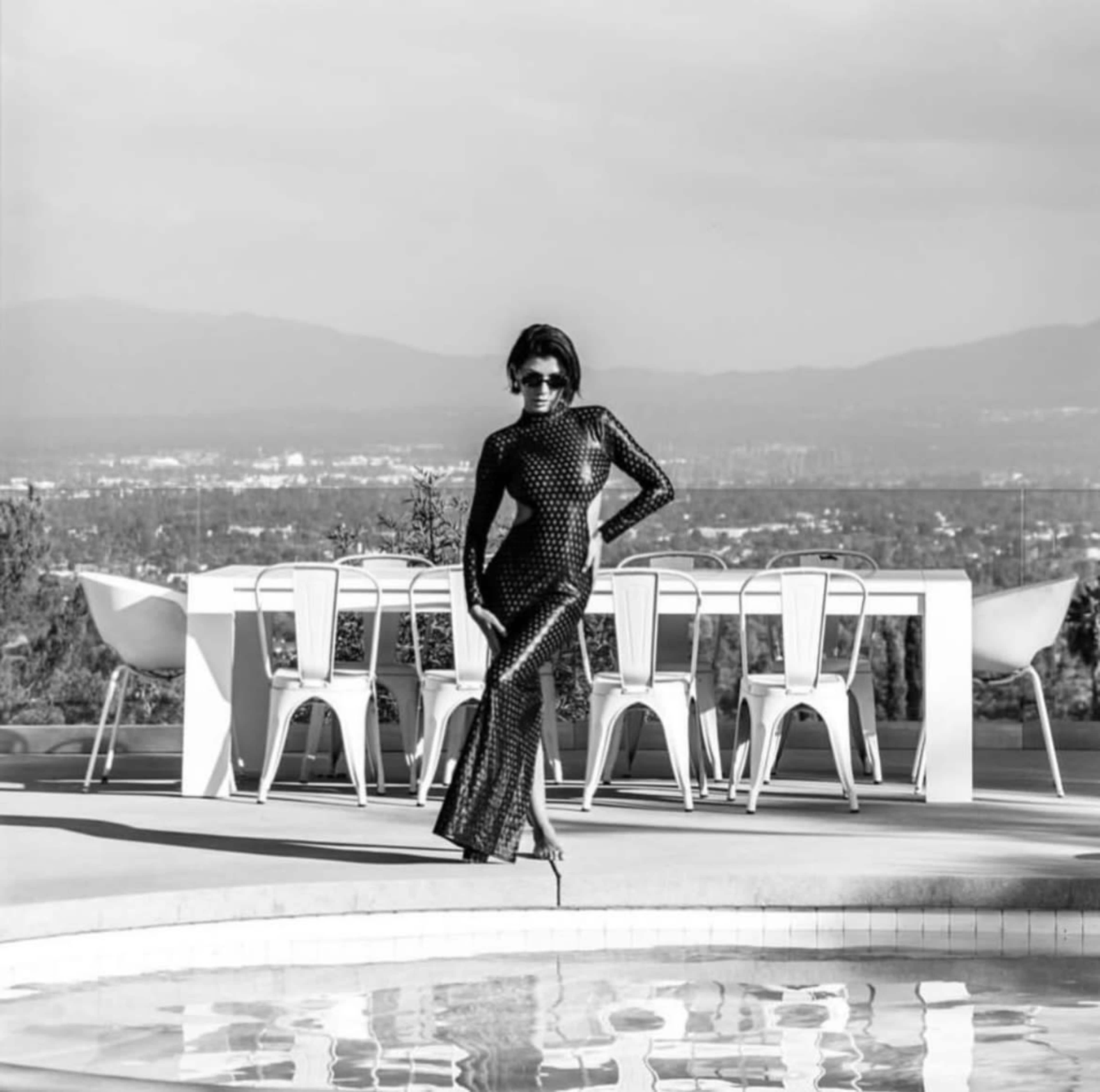 A woman in a form-fitting, patterned dress stands beside a pool in front of a modern outdoor dining setting with white chairs and a table.