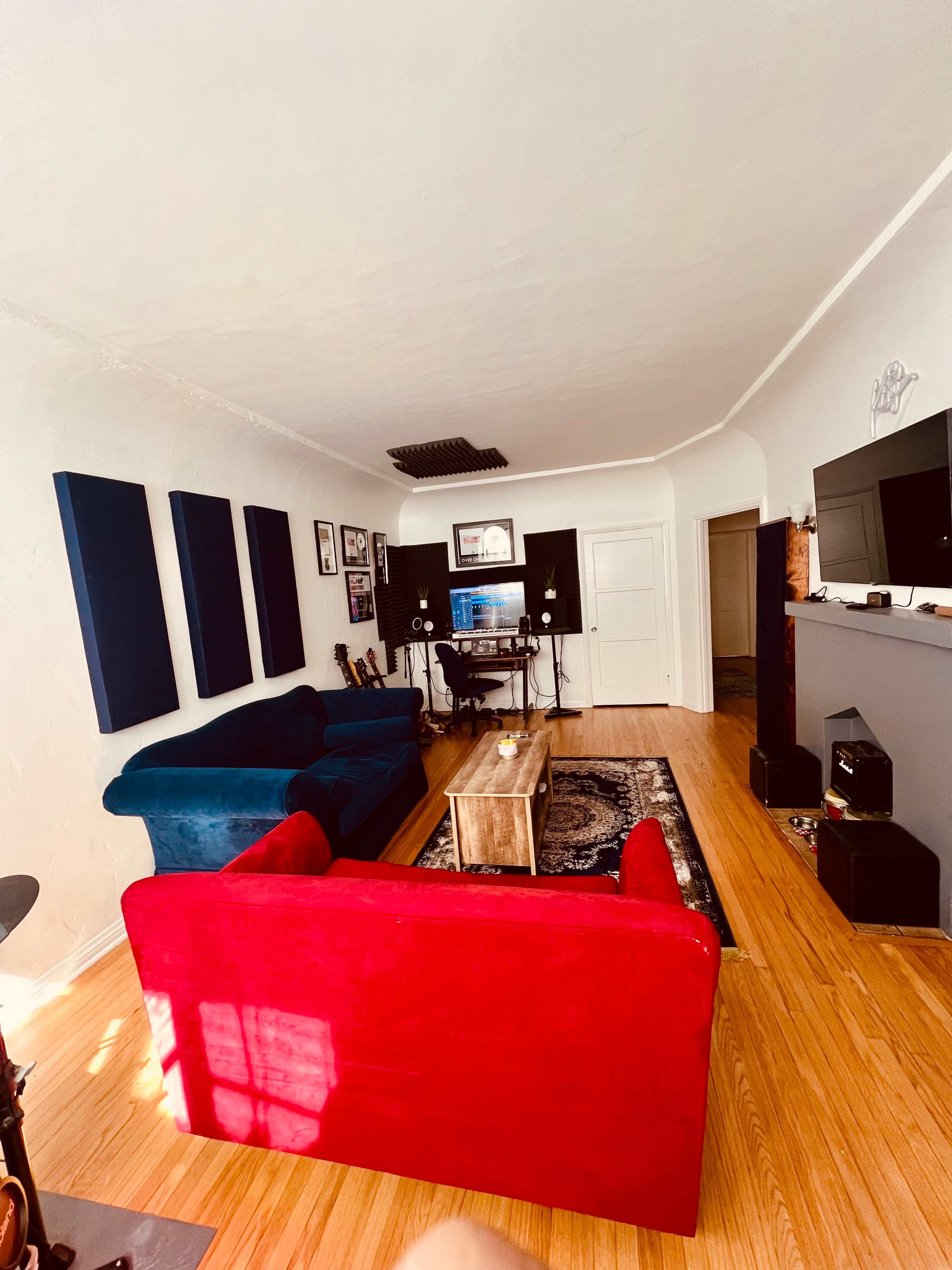 A living room with a red couch and a blue couch, a wooden coffee table, and a workspace featuring black sound panels and a computer setup against a white wall.