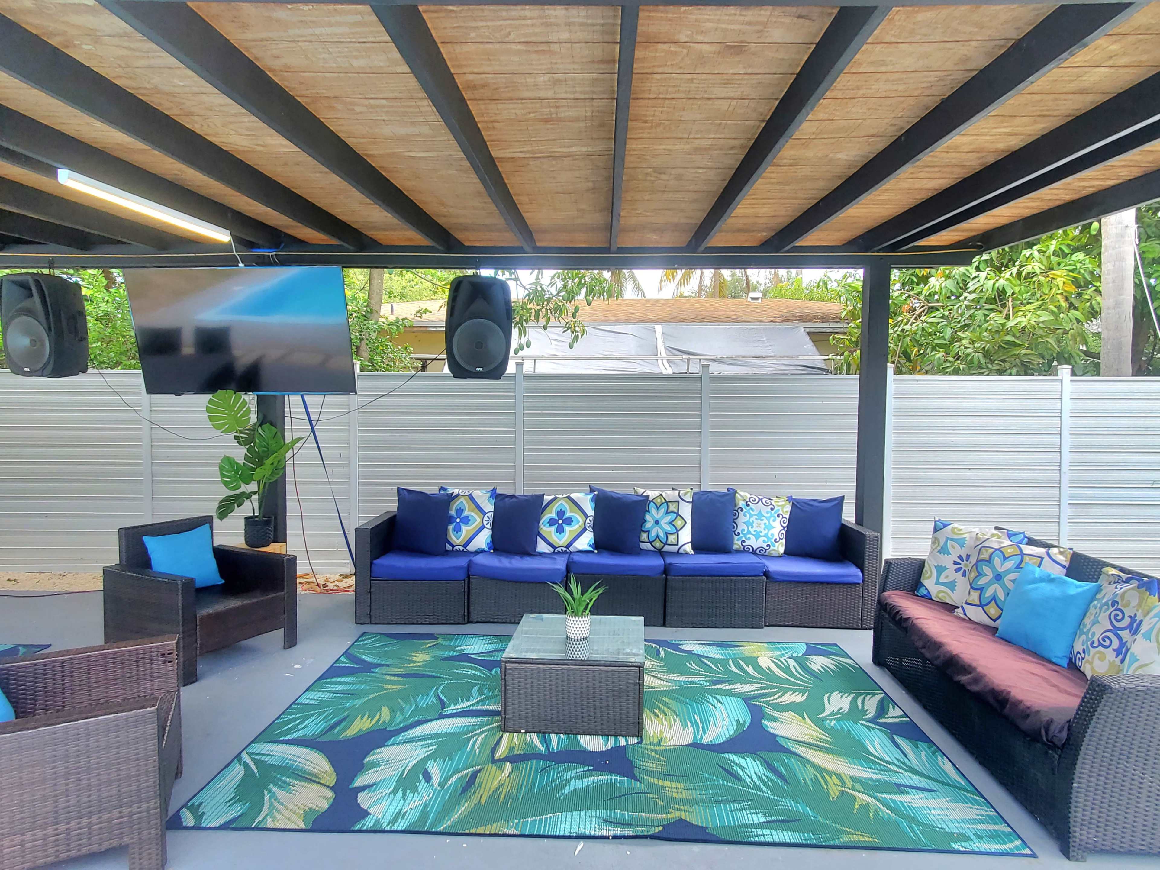 A covered outdoor patio features dark wicker furniture with colorful cushions arranged around a large area rug, along with a television mounted on the wall.