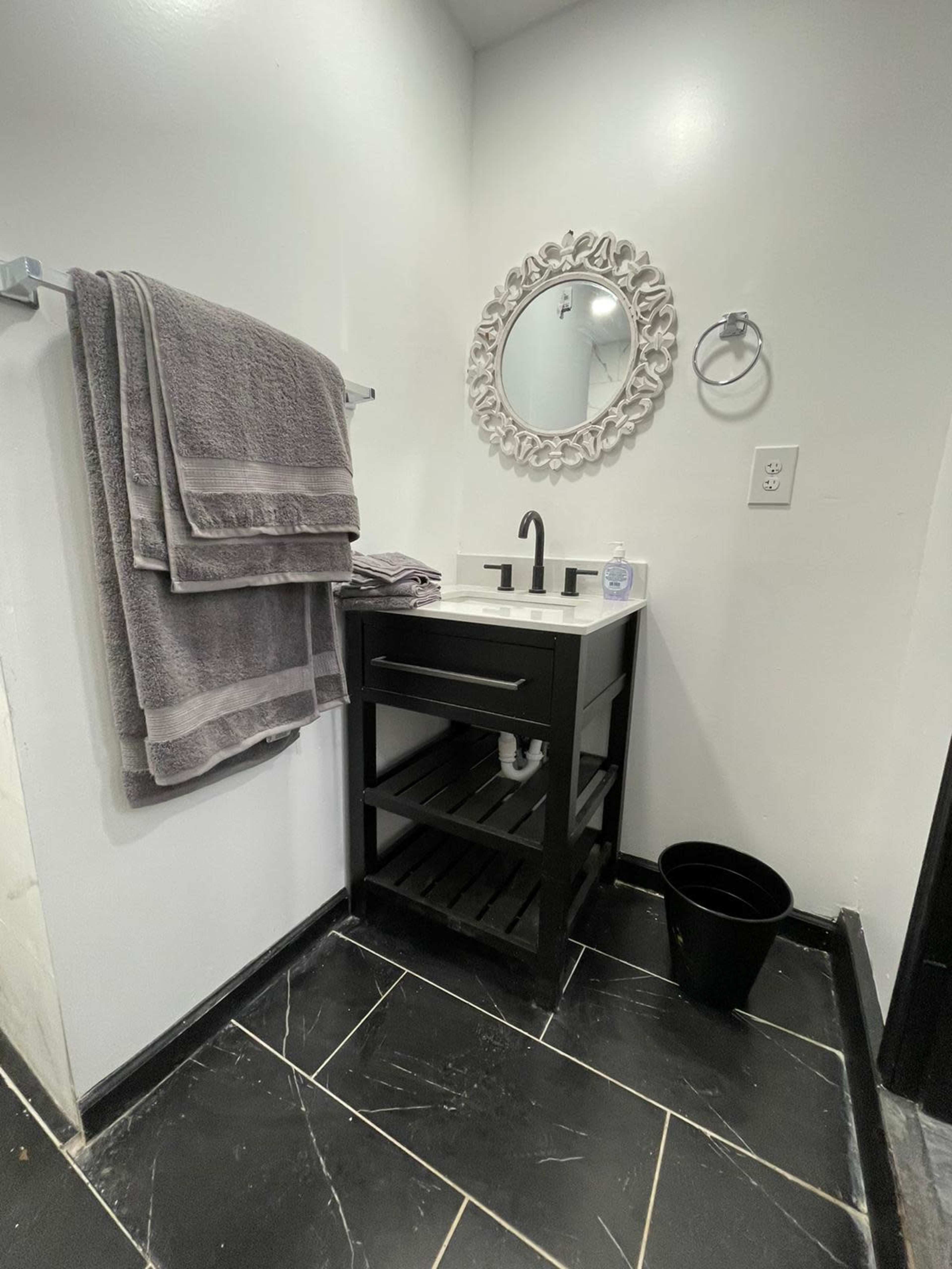 A small bathroom features a black vanity with a sink, a round mirror, and neatly hung gray towels on a towel rack, with black tiles on the floor.