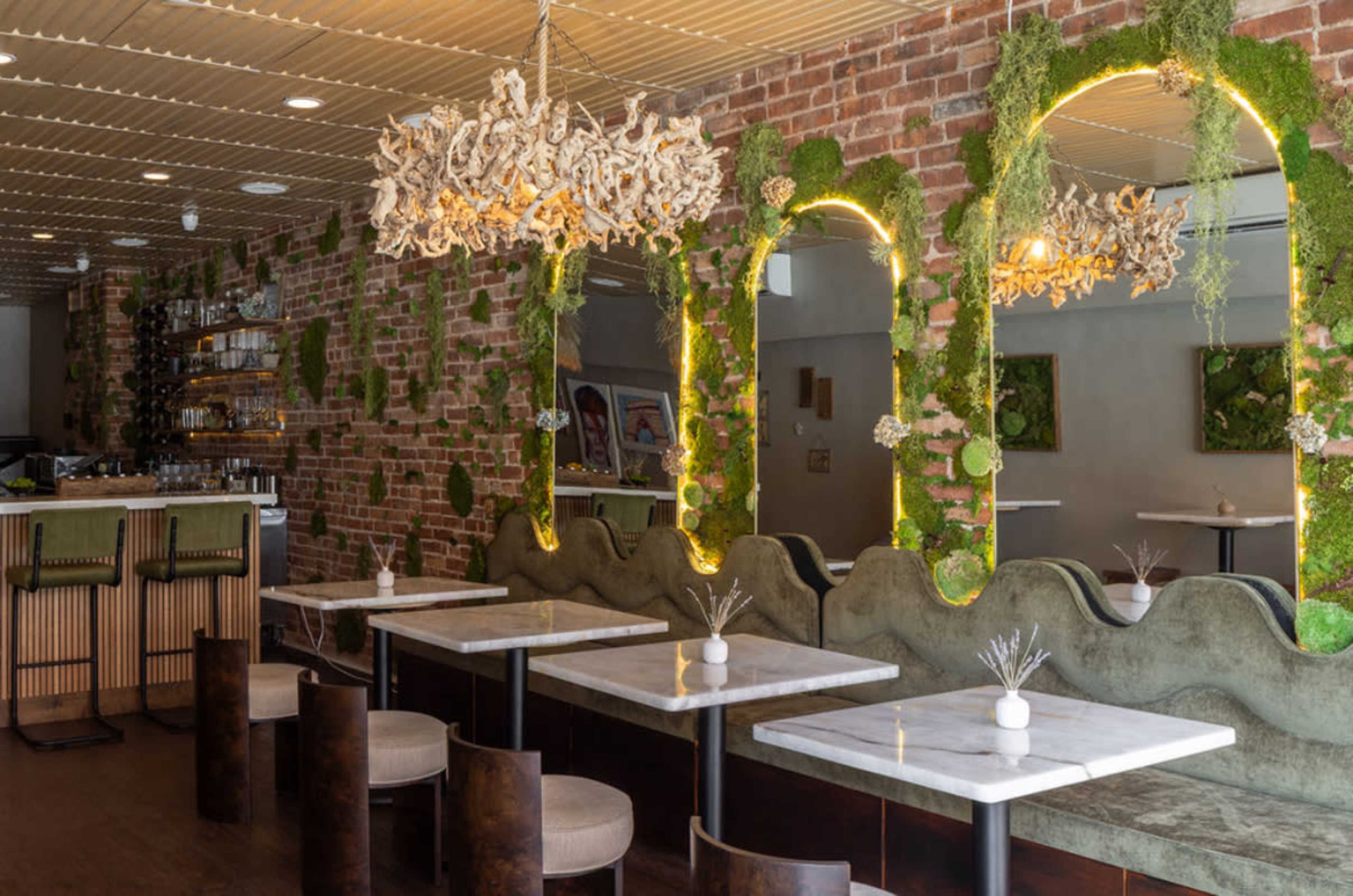 The image shows a modern restaurant interior with brick walls adorned with greenery, featuring marble-topped tables and unique lighting fixtures.