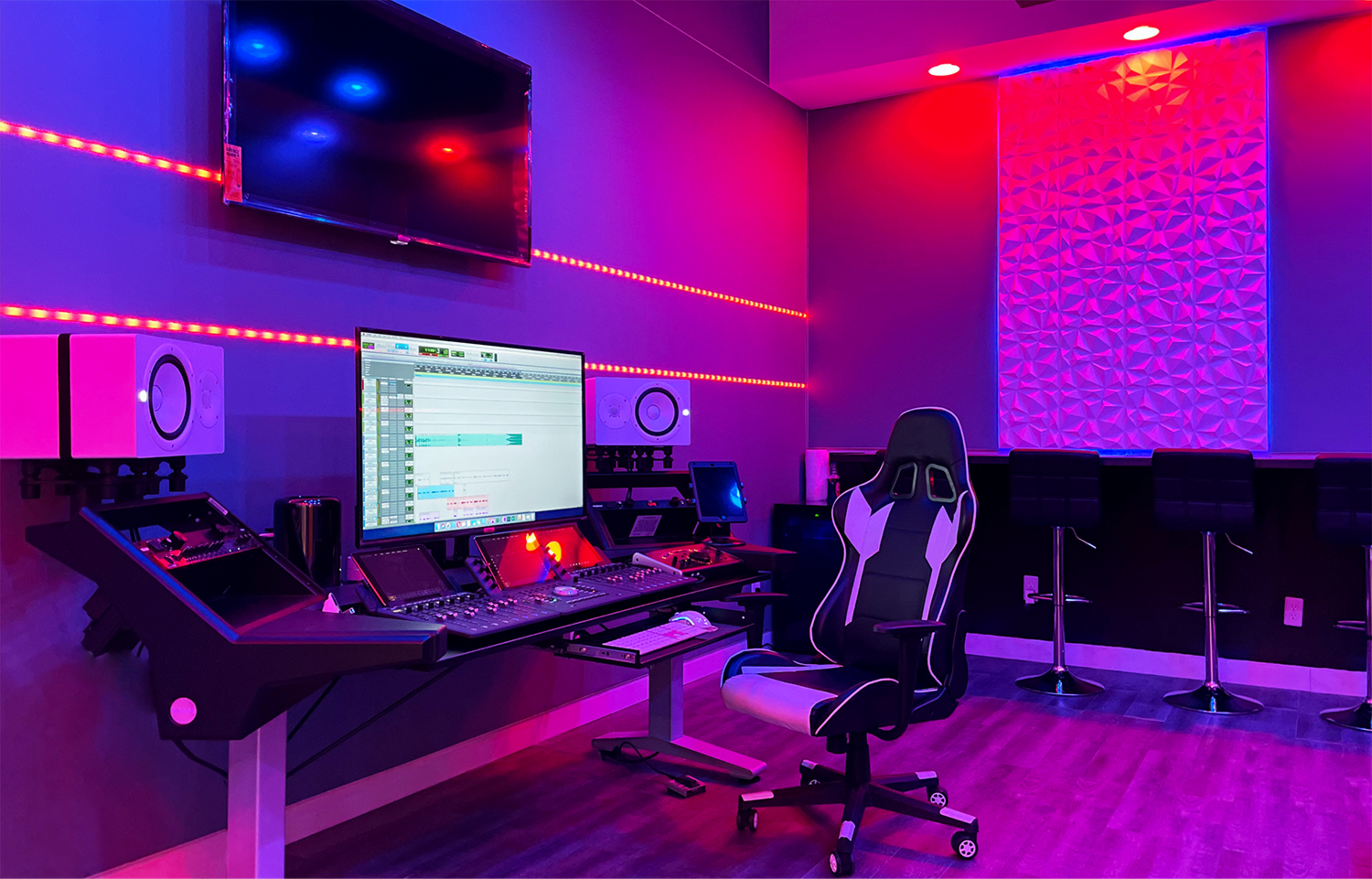 A modern recording studio features a large computer monitor, a sleek mixing desk with audio equipment, and colorful LED lighting.