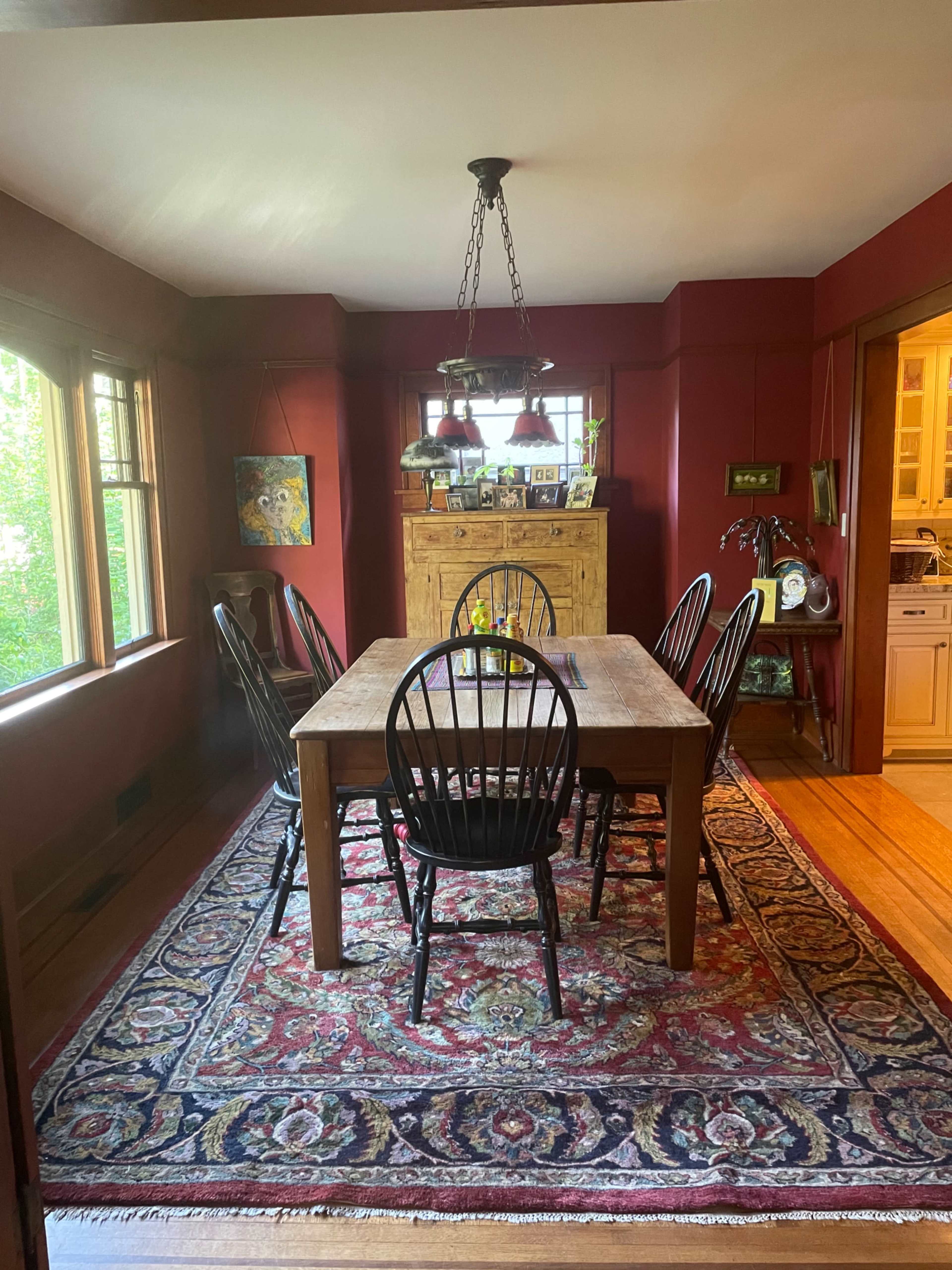 A dining room features a wooden table surrounded by black chairs, with large windows and a patterned rug on the floor.
