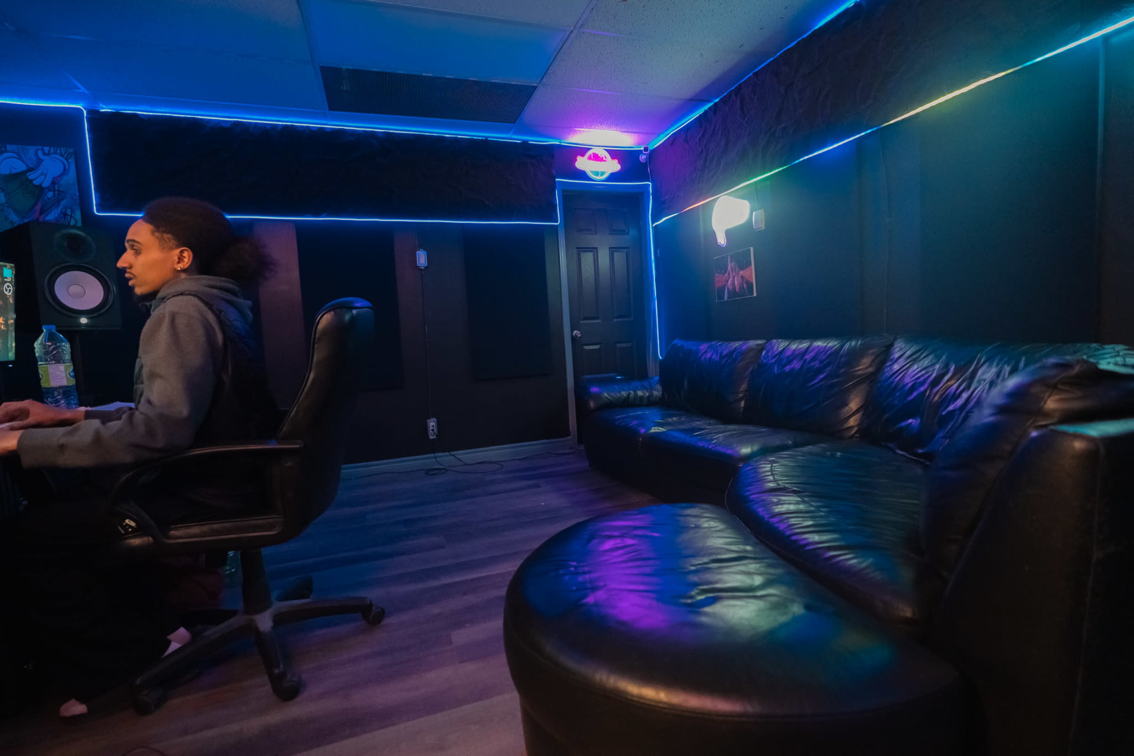 A person sits at a desk in a recording studio with dark walls and neon lighting, while a curved leather sofa is positioned nearby.