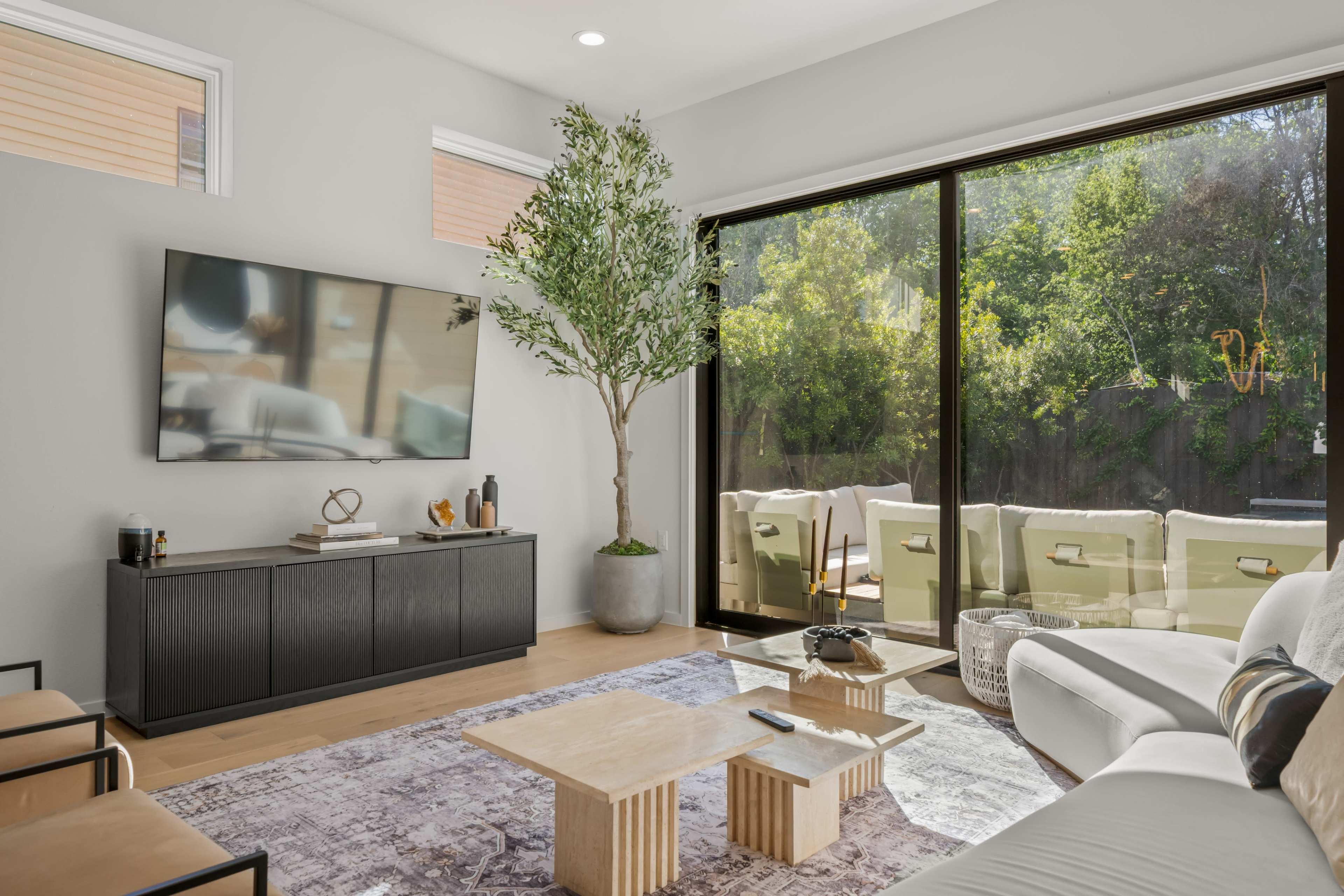 A modern living room features a large television mounted on a wall, a stylish coffee table, and a potted tree, with large glass doors leading to a patio surrounded by greenery.