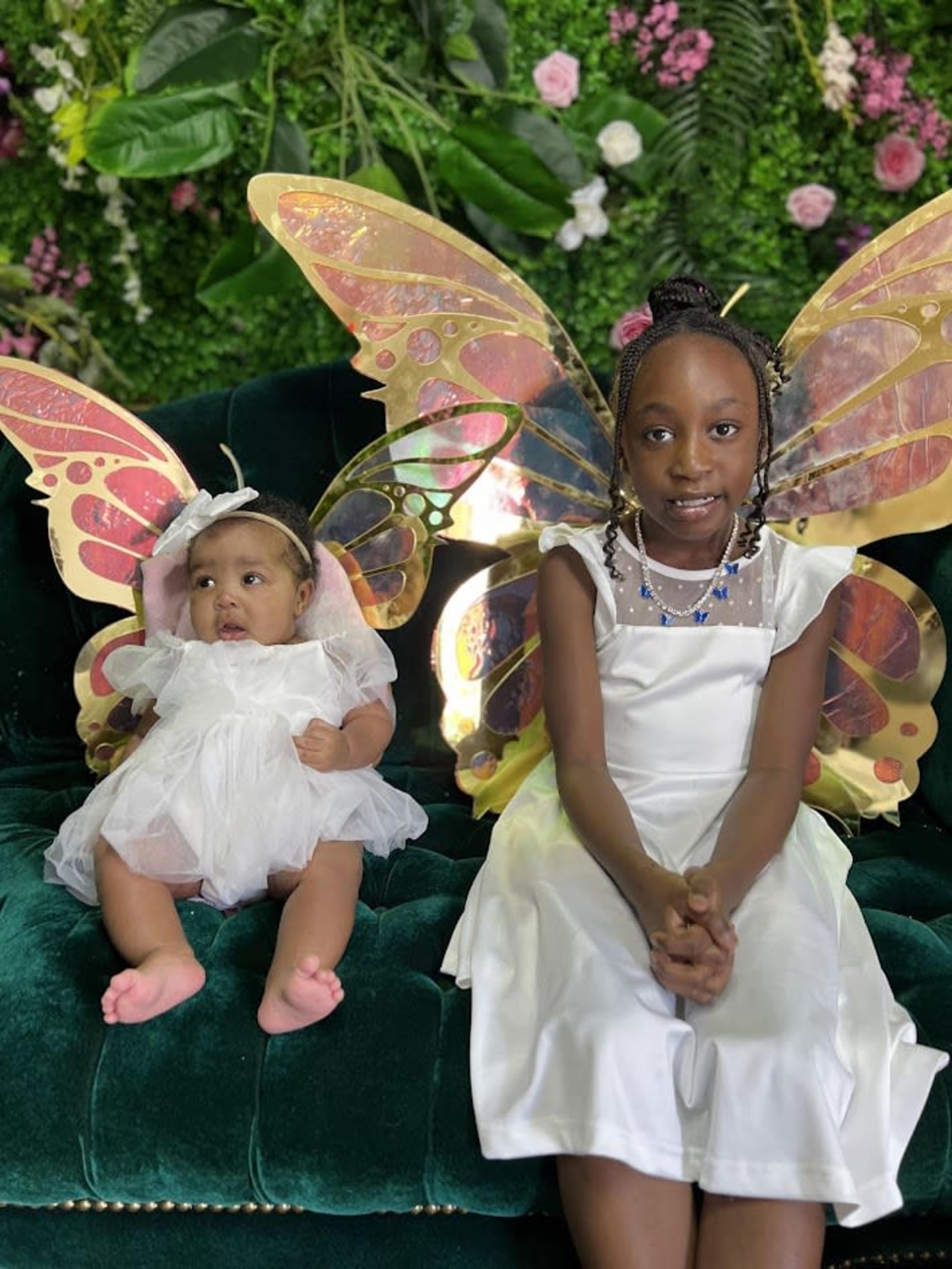 Two children dressed in white, with large colorful butterfly wings, are sitting on a green couch in front of a floral backdrop.