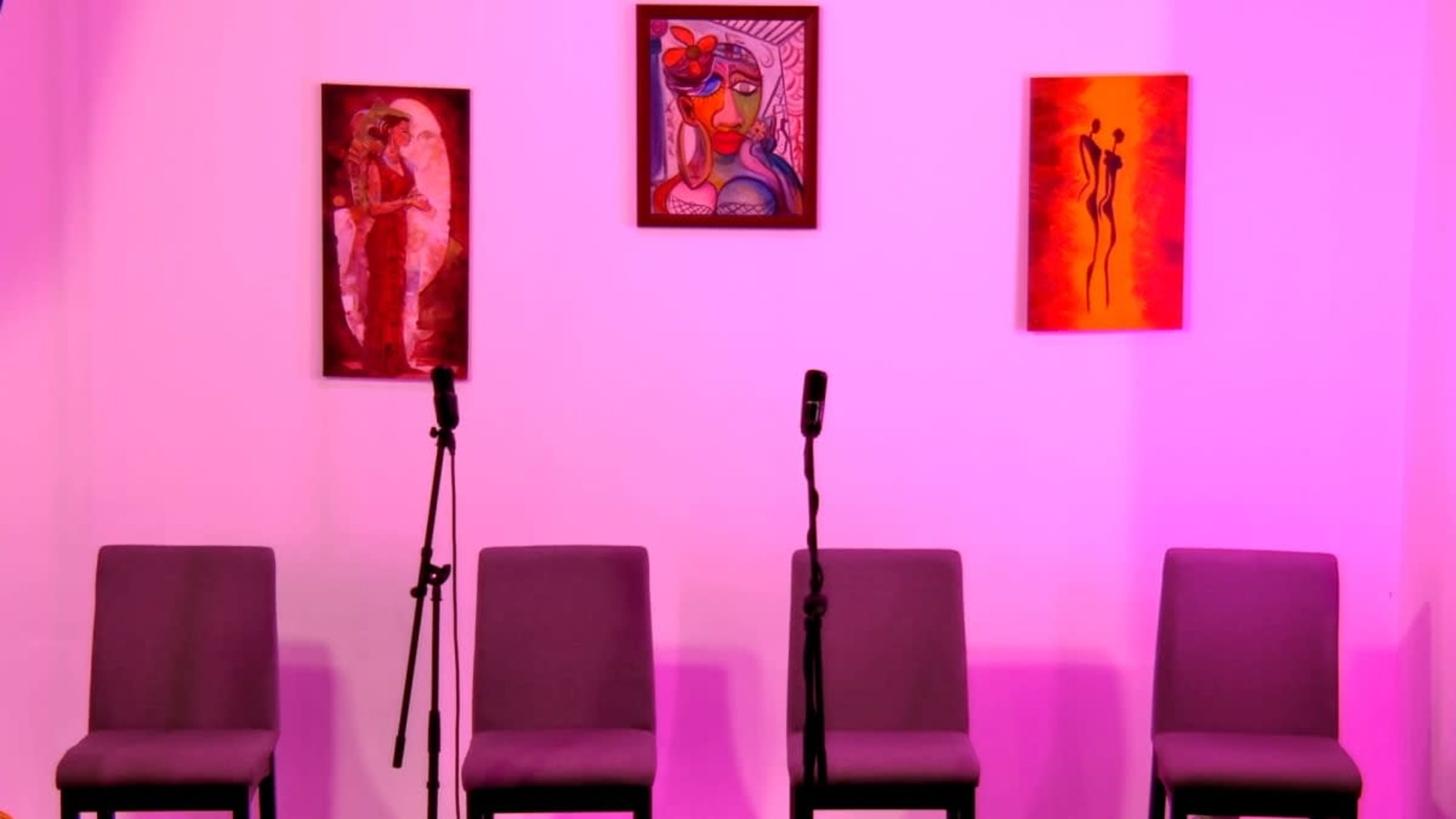 The image shows four empty chairs arranged in front of two microphones against a backdrop of colorful artwork on a pink wall.