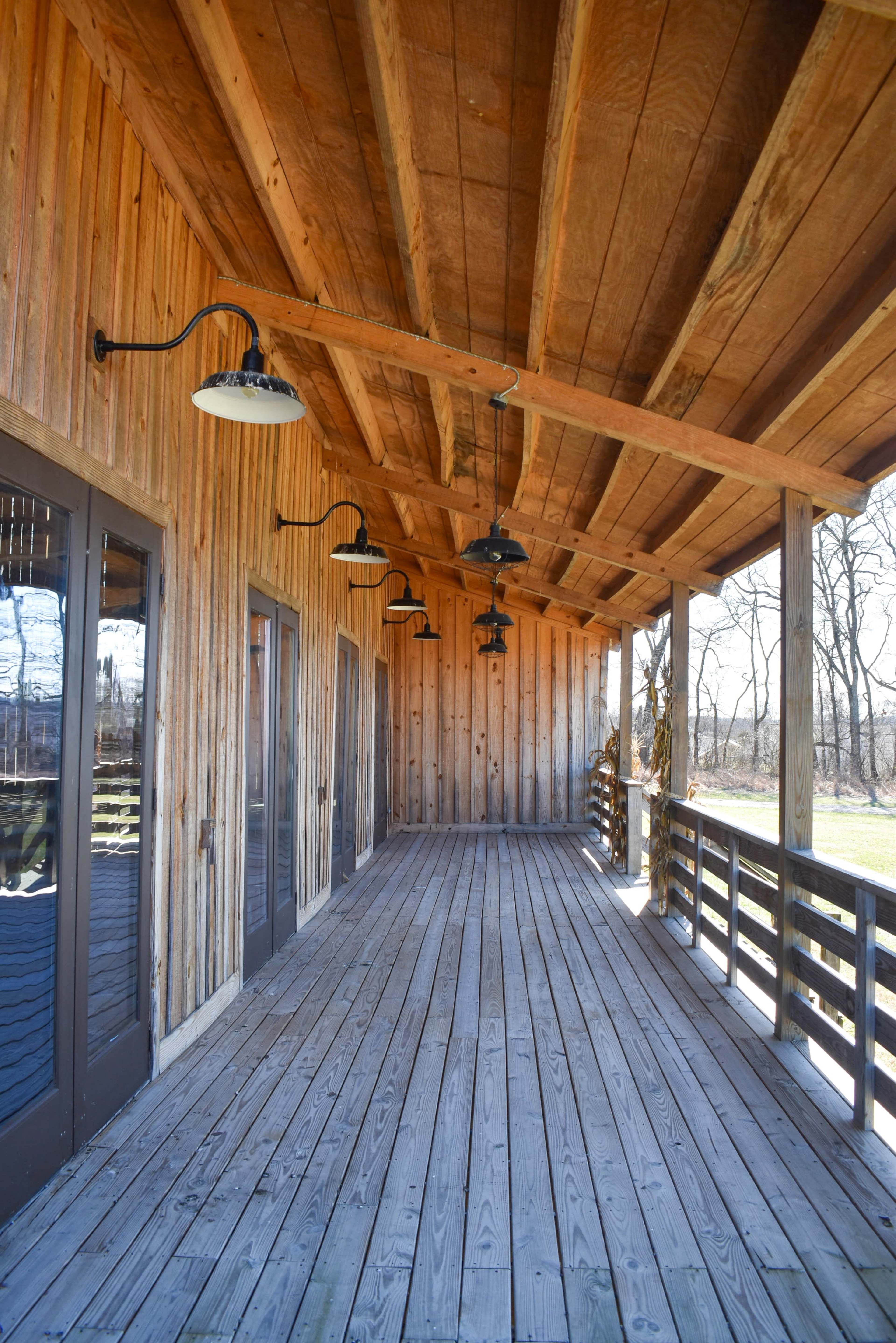 The image shows a wooden porch with a slatted floor and overhead lights, leading to double doors that open to a rustic exterior.