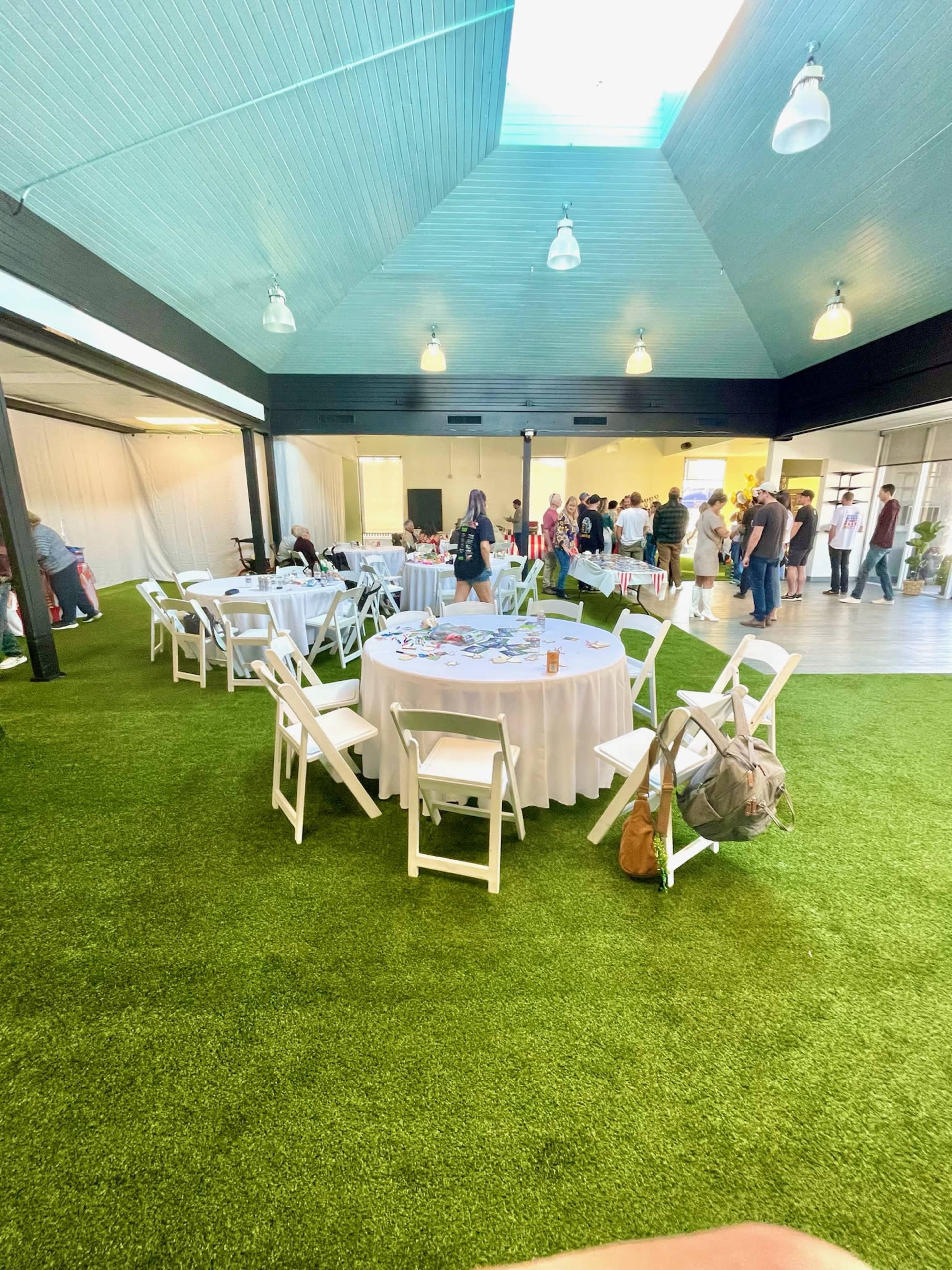 The image shows an event space with several round tables covered in white tablecloths, surrounded by white chairs, and a crowd of people engaging in activity in the background.