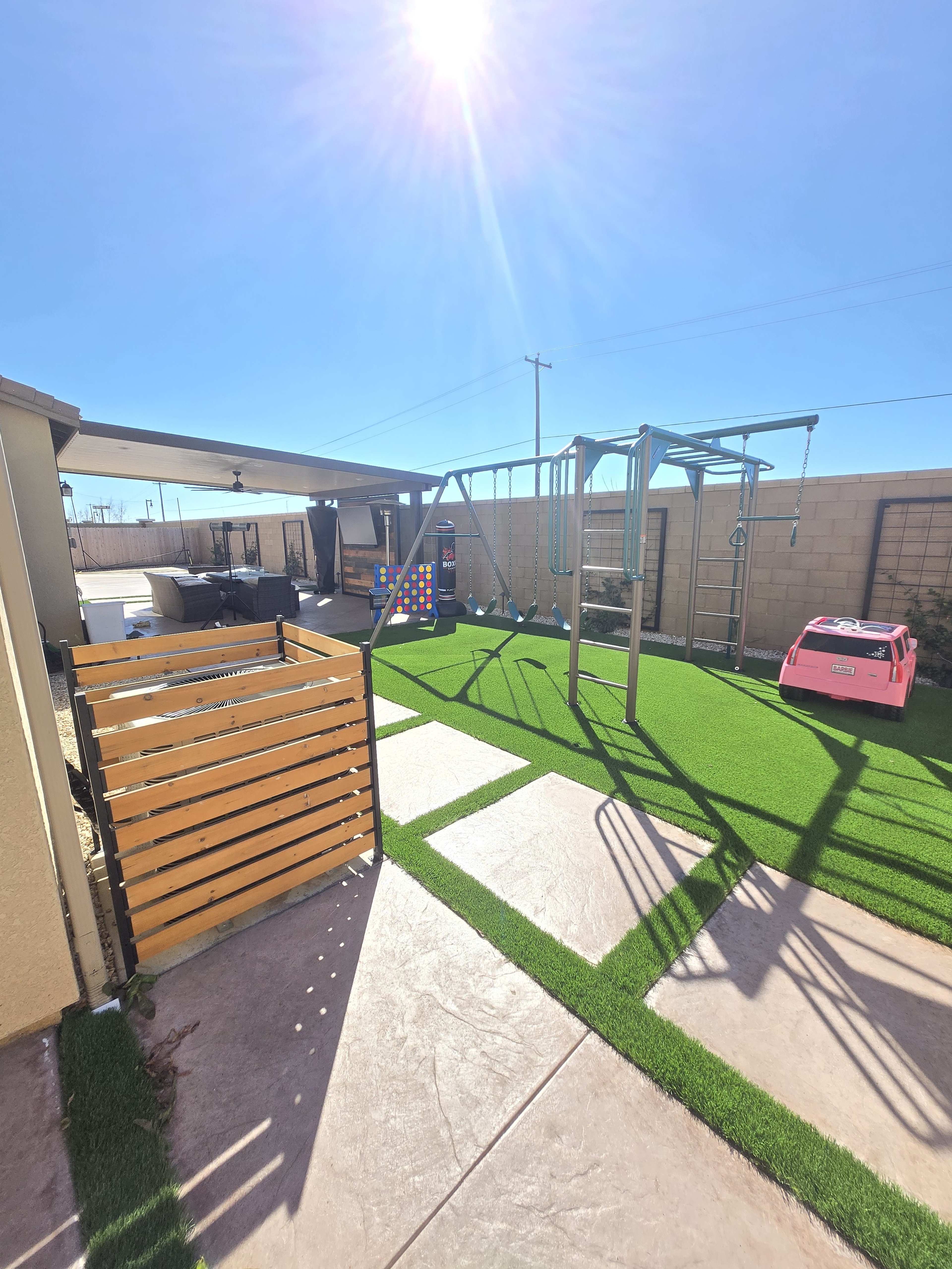 The image shows a backyard with a playset, swing, and a pink toy car on artificial grass, surrounded by a wall and a shaded patio area.