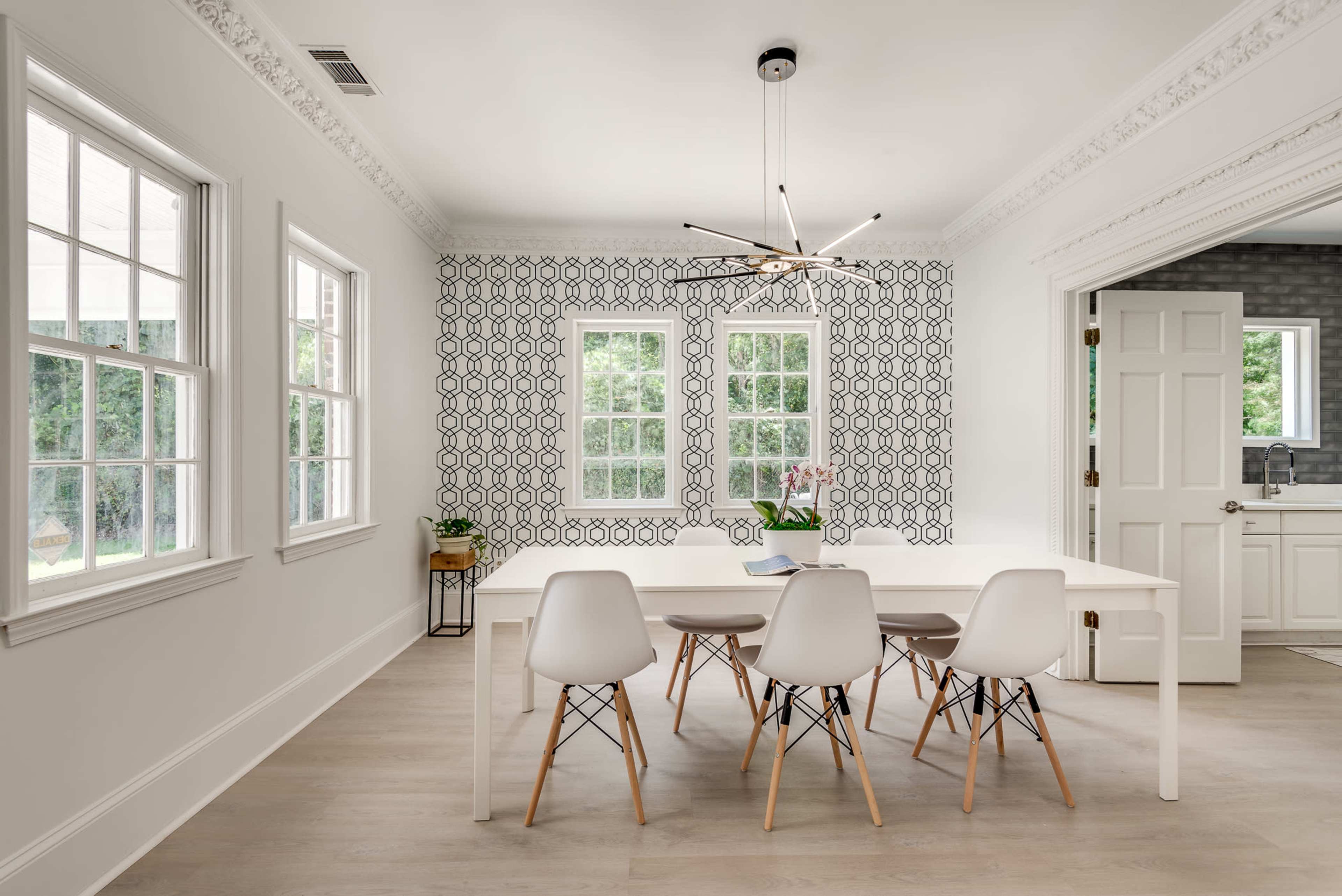 A bright dining room features a white table with six chairs, surrounded by patterned wallpaper and large windows.