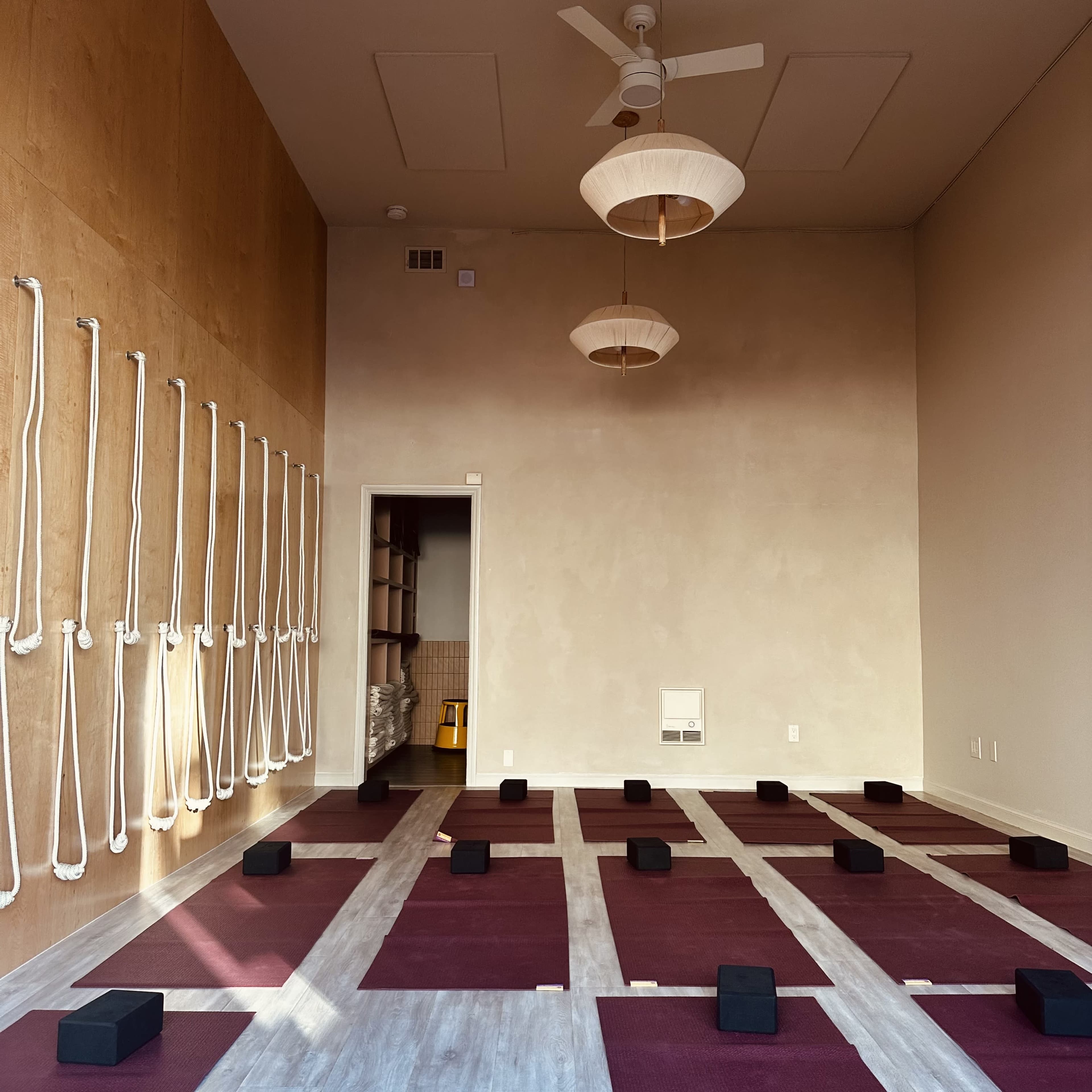 The image shows an empty yoga studio with maroon mats, black yoga blocks arranged on the floor, and ropes mounted on the wall.