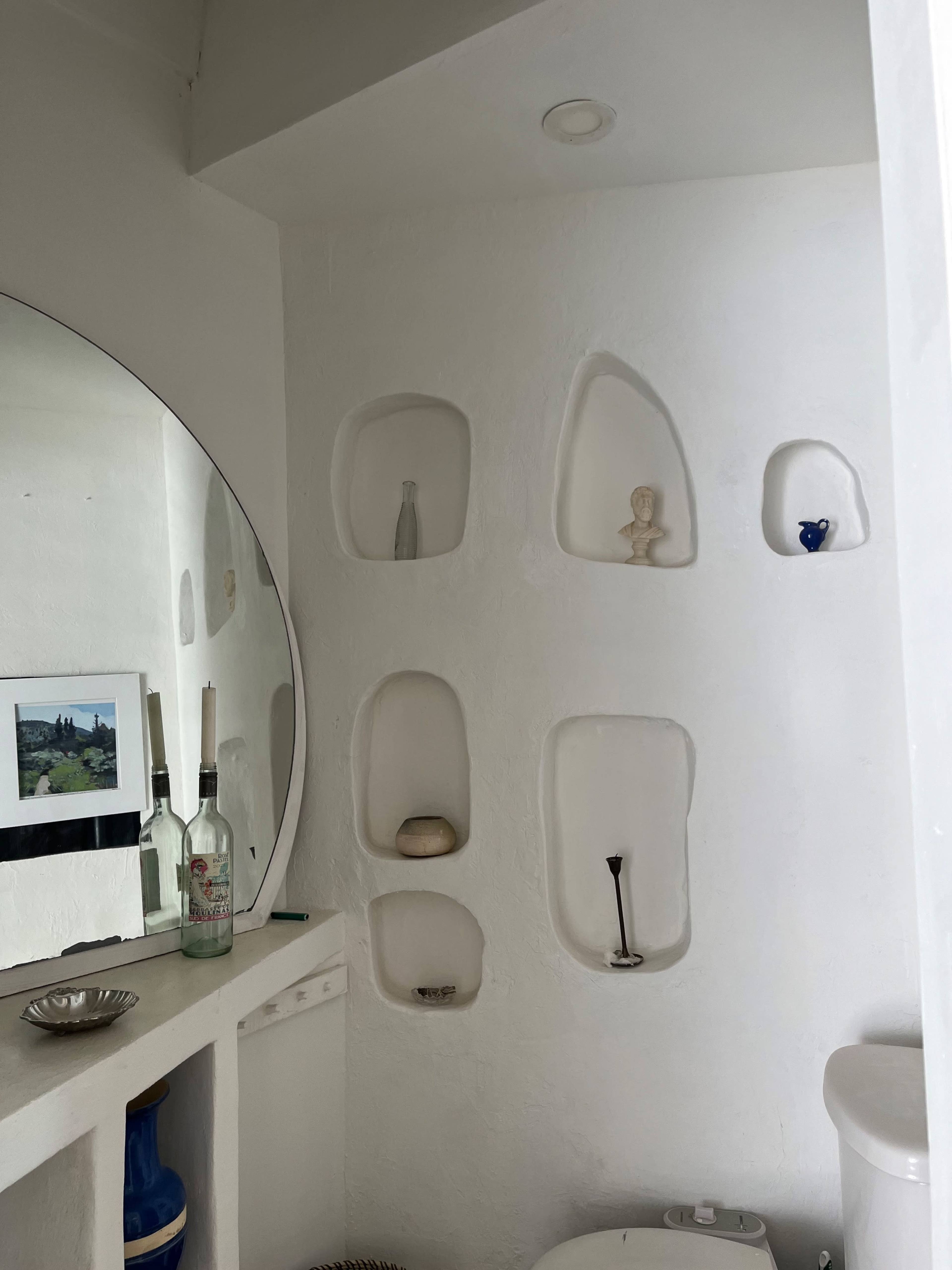 A small bathroom with a round mirror, white walls featuring niche shelves, and minimalist decor items.