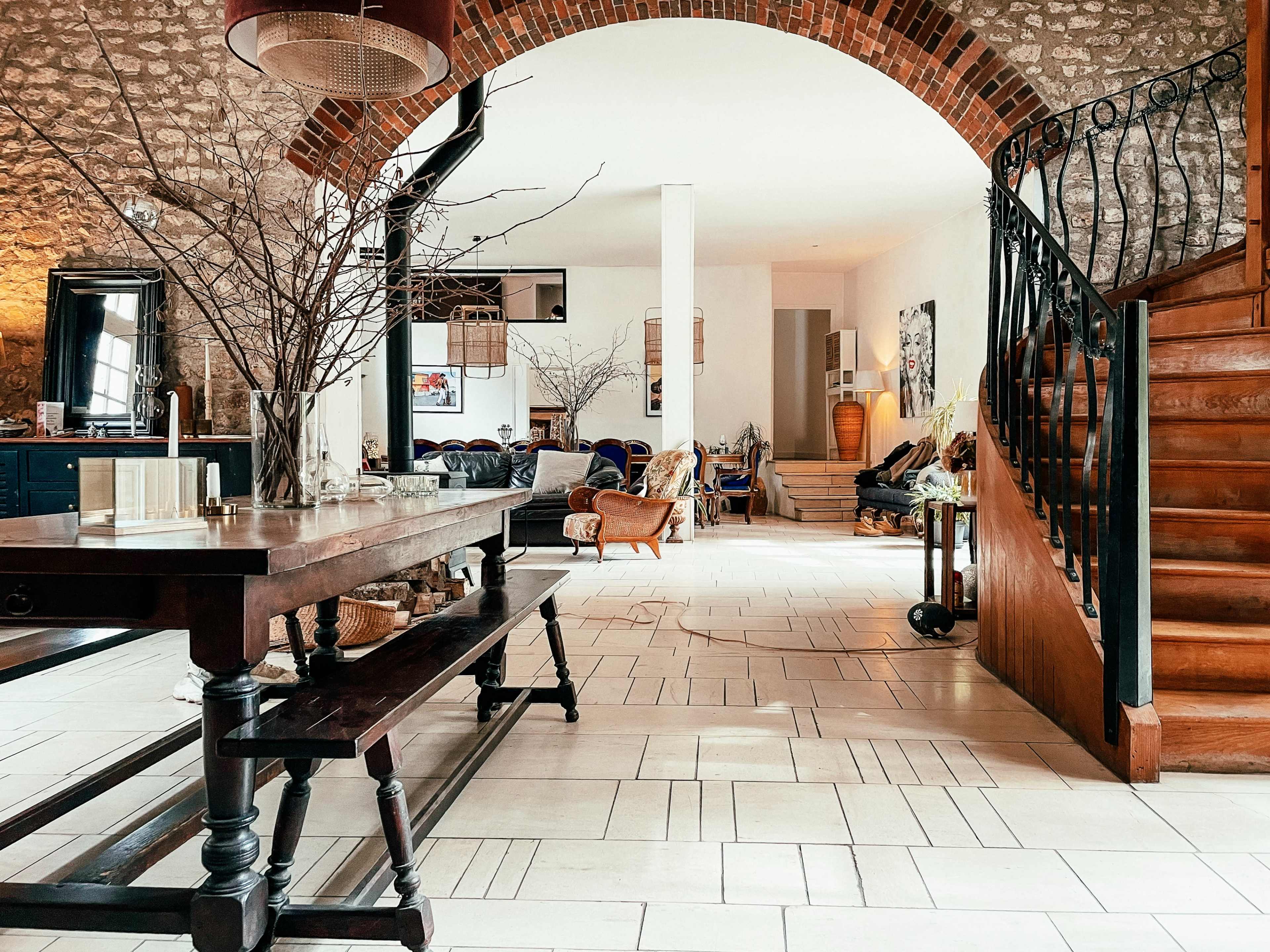 The image depicts a spacious interior with a wooden dining table, a staircase, and various seating areas, characterized by stone walls and large windows.