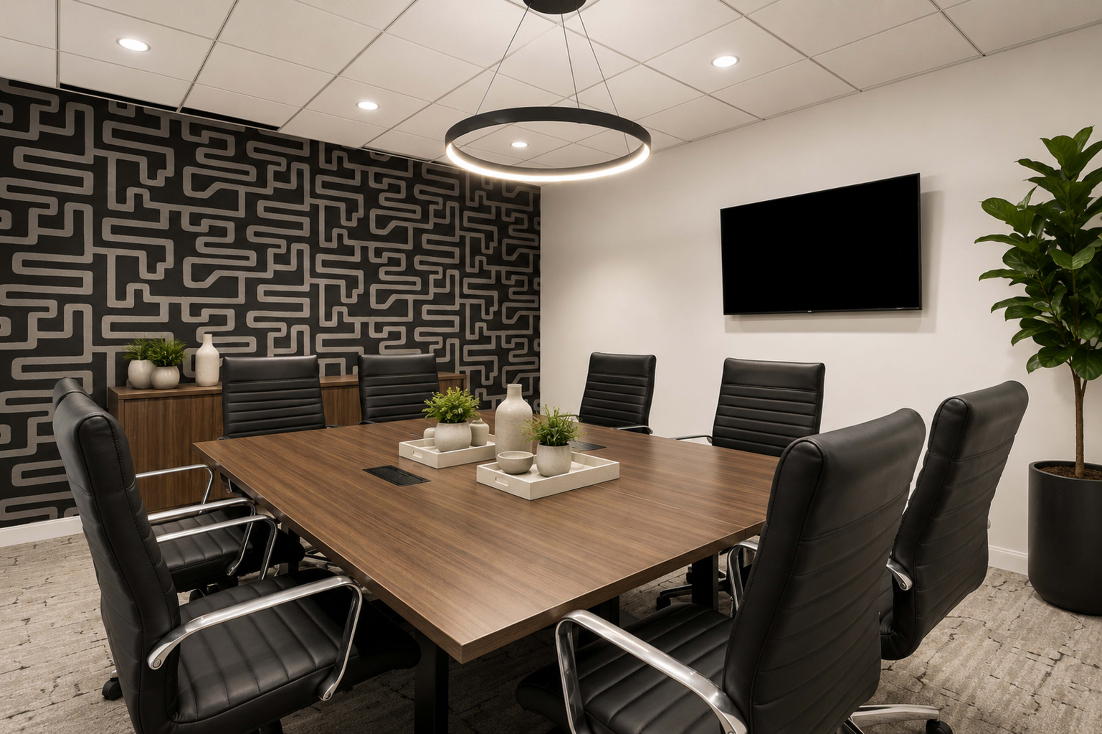 The image shows a modern conference room with a large wooden table surrounded by black leather chairs, a patterned rear wall, and a television mounted on the wall.
