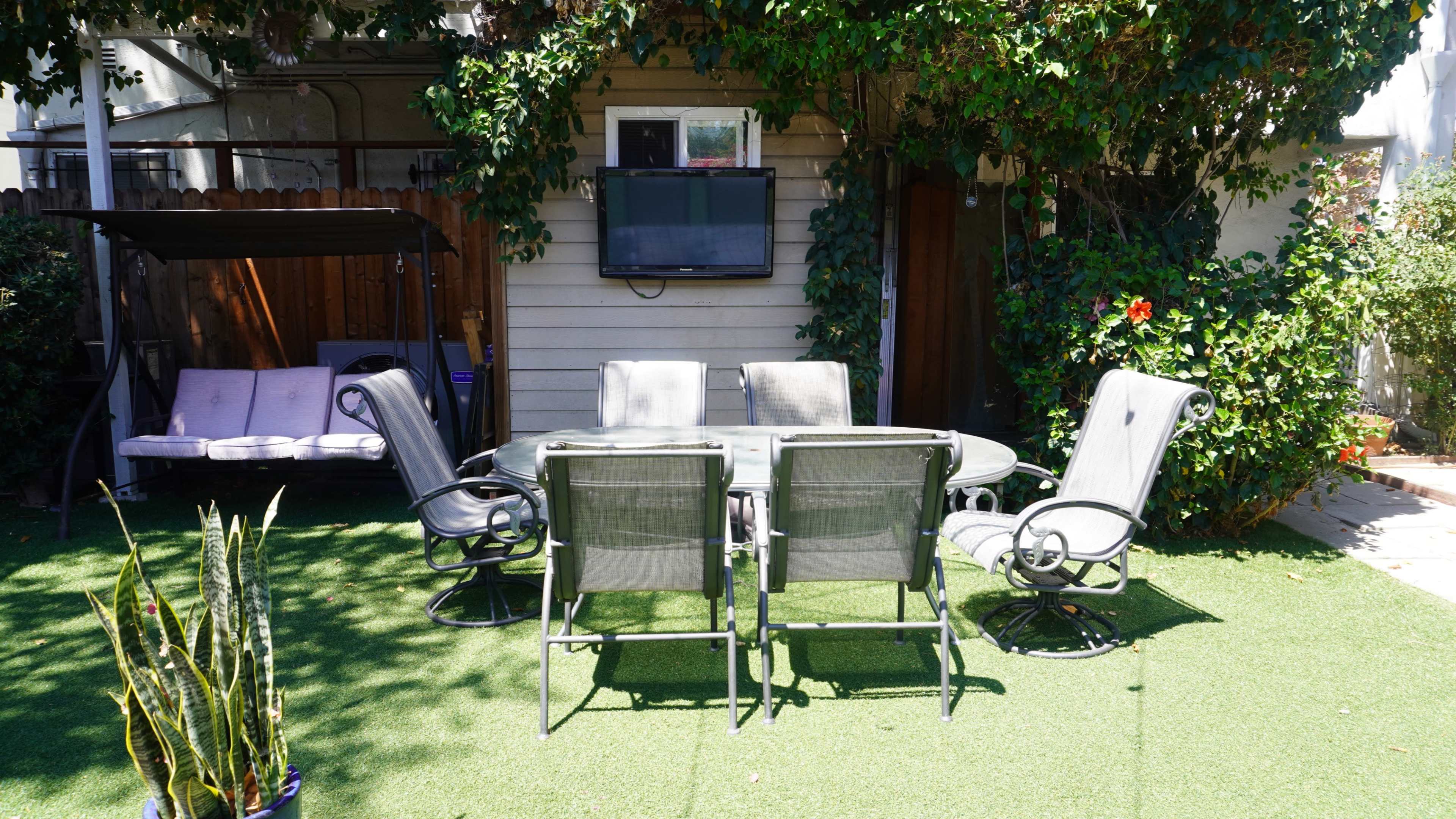 A patio area features a glass dining table surrounded by six chairs, with a swing and a mounted television visible in the background.