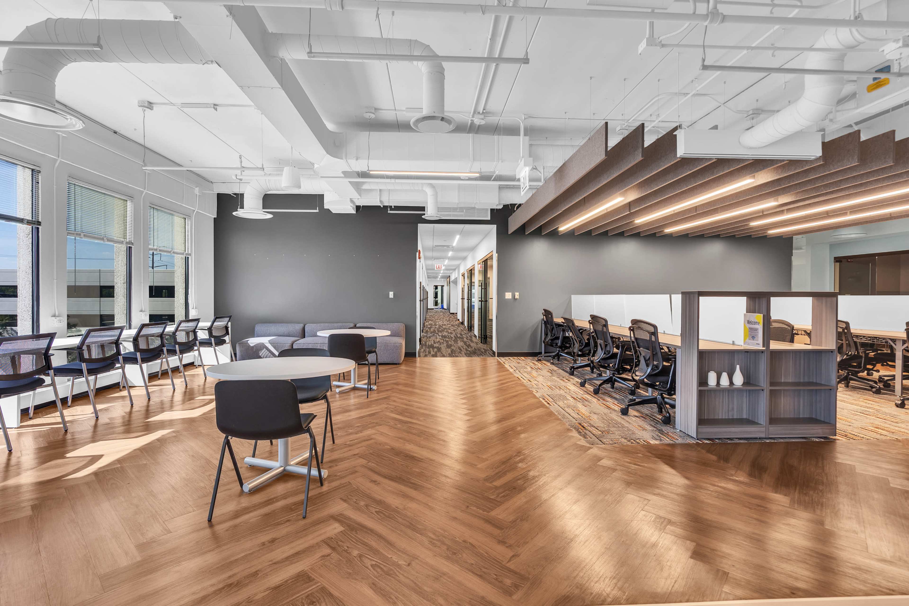 The image shows a modern office space with wooden flooring, desks arranged in a semi-open layout, and large windows allowing natural light.