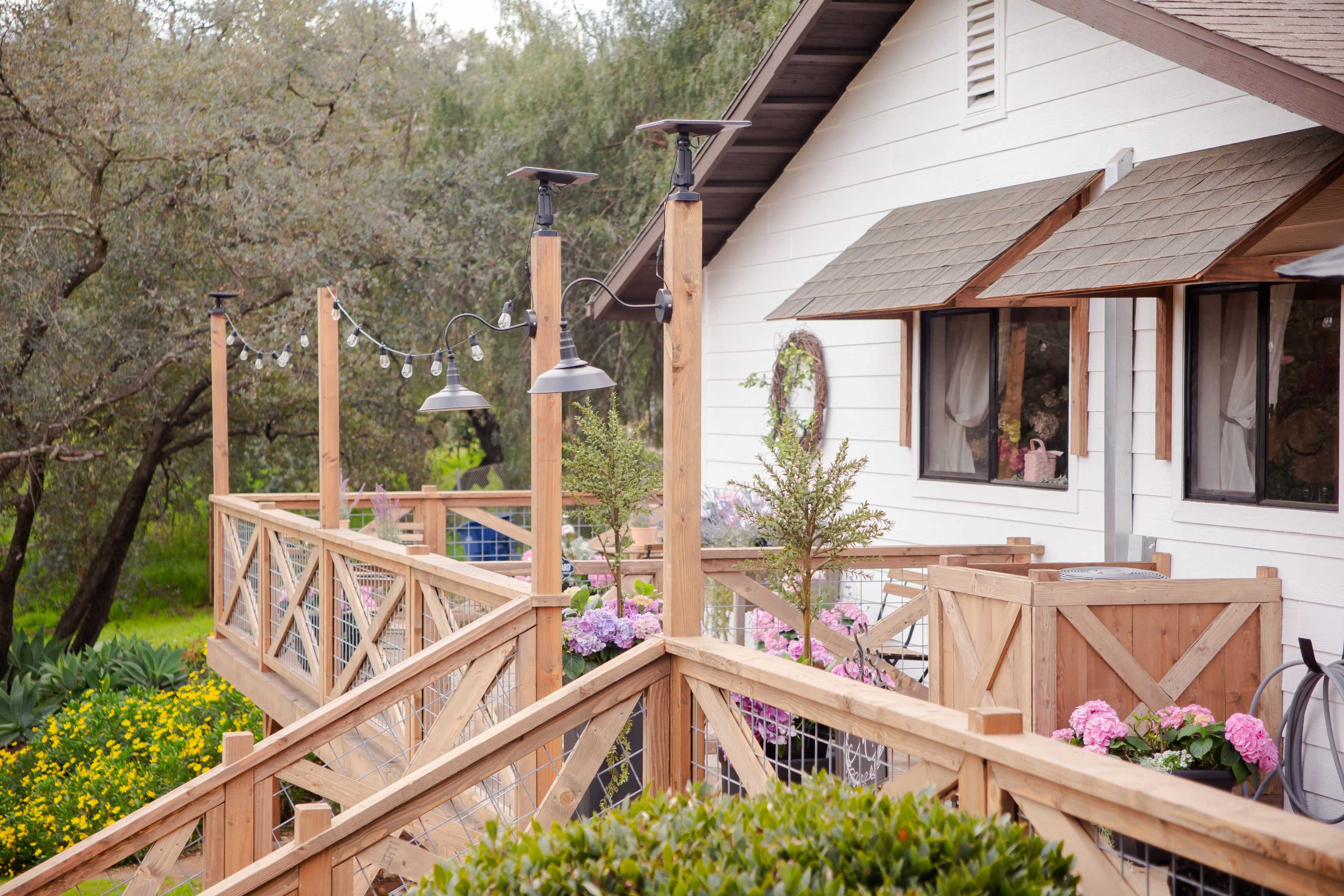 A wooden deck with a railing adorned with plants and lights leads to a cozy white house surrounded by greenery and flowers.