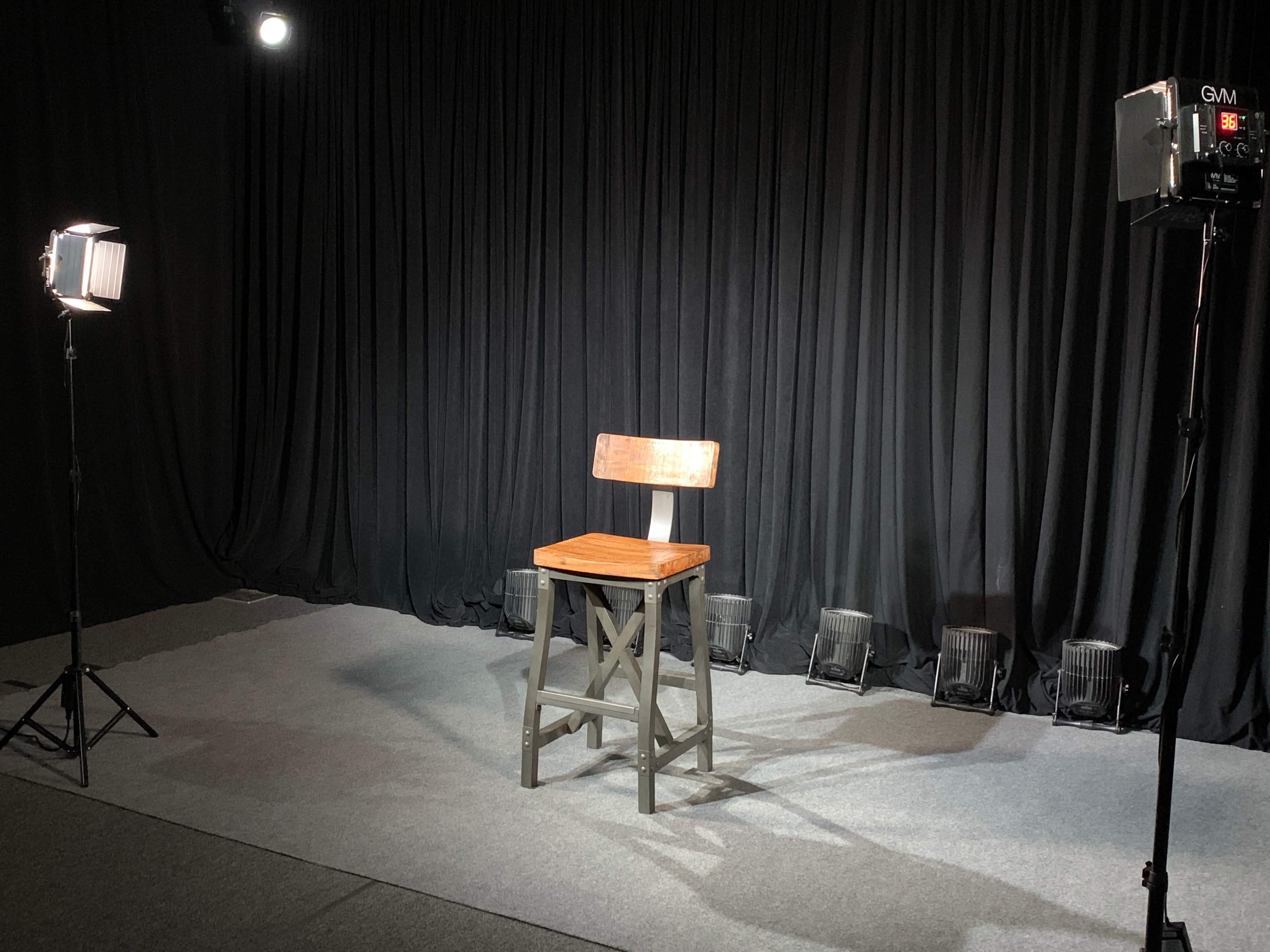 A wooden stool is positioned on a gray carpeted area, surrounded by two lights and a backdrop of black curtains.