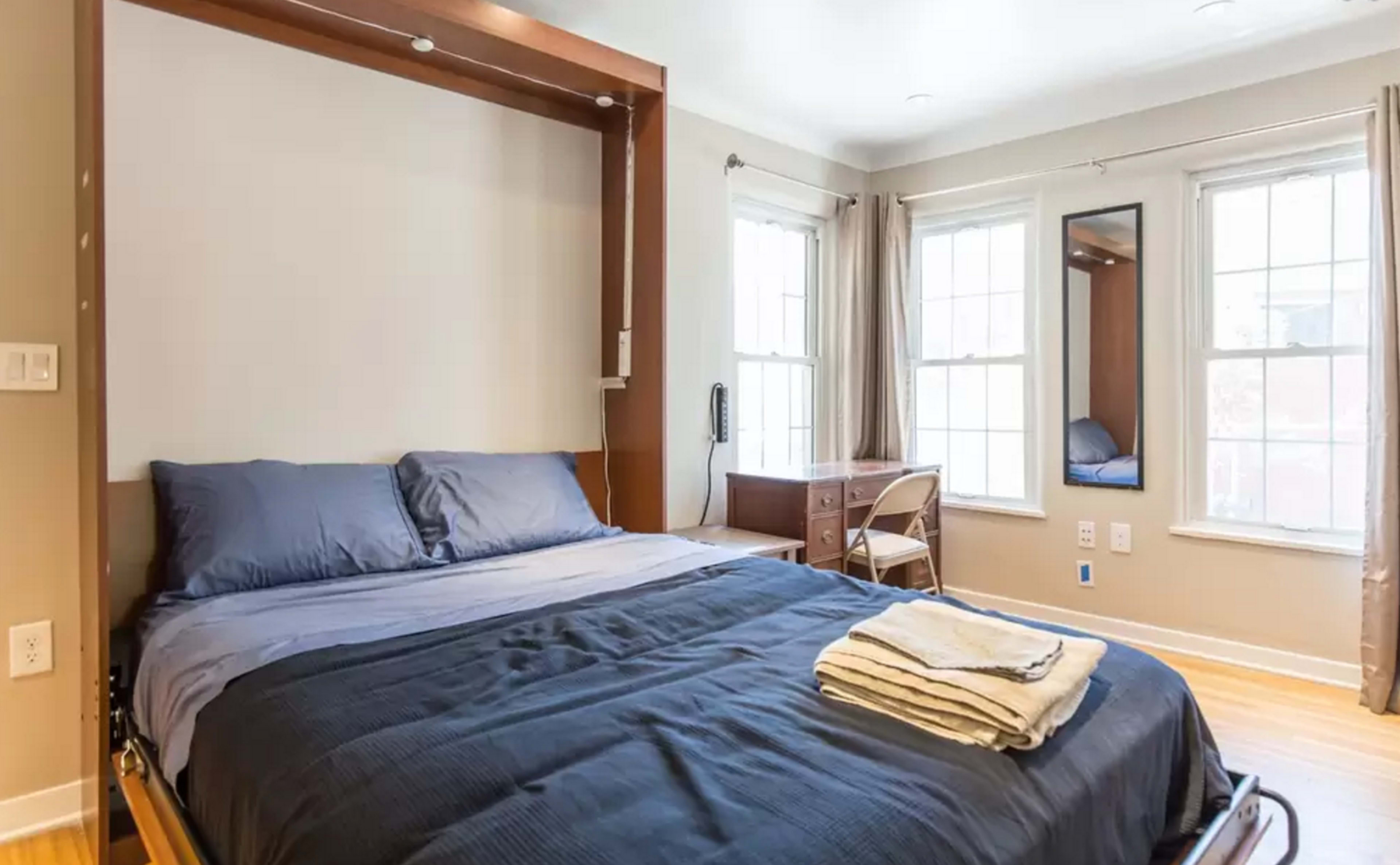 A neatly arranged bedroom with a foldable wall bed, accompanied by a small desk and two large windows allowing natural light.