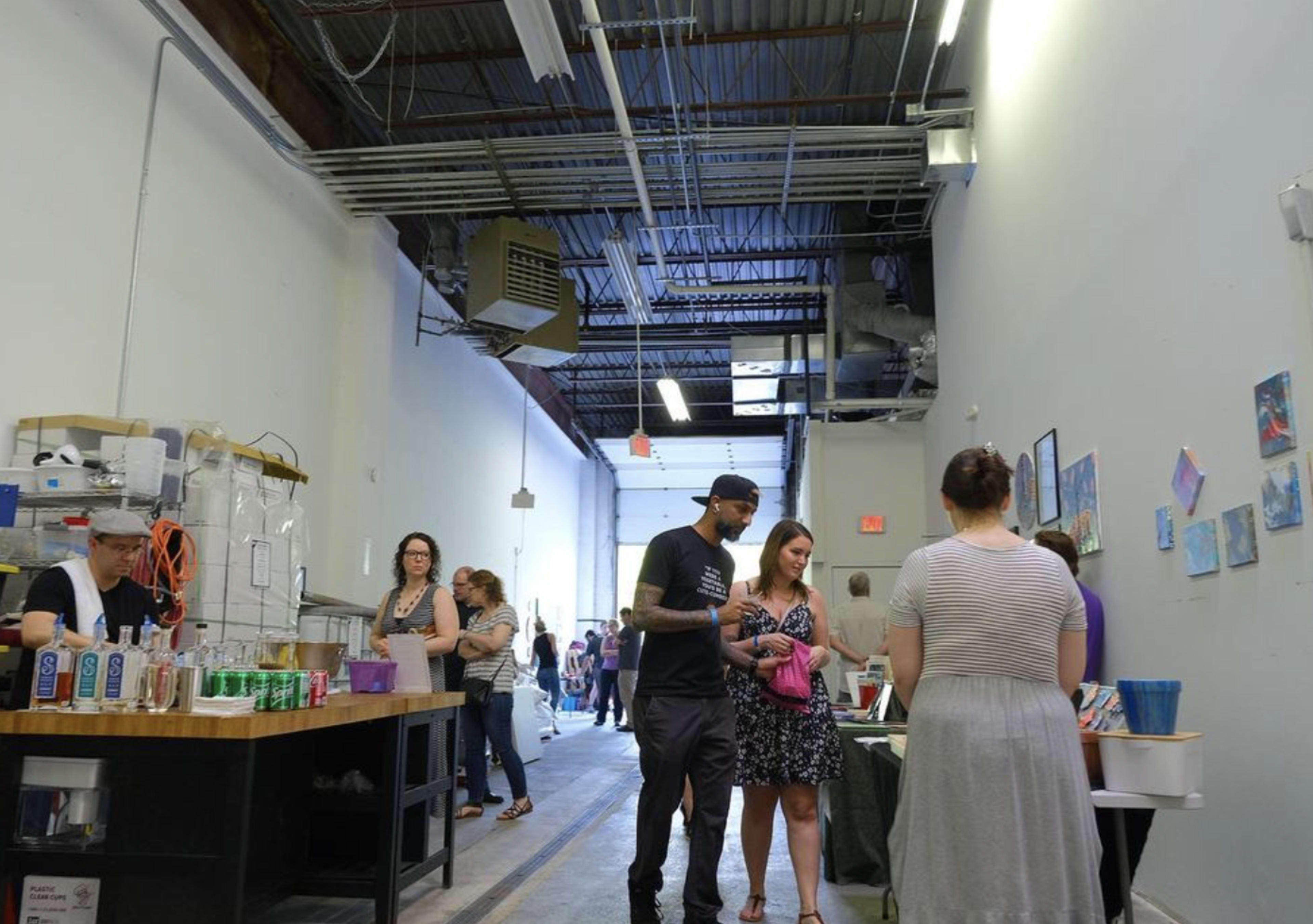 A group of people engages in conversation and browsing along an open corridor, with tables displaying various items and a counter serving beverages on one side.