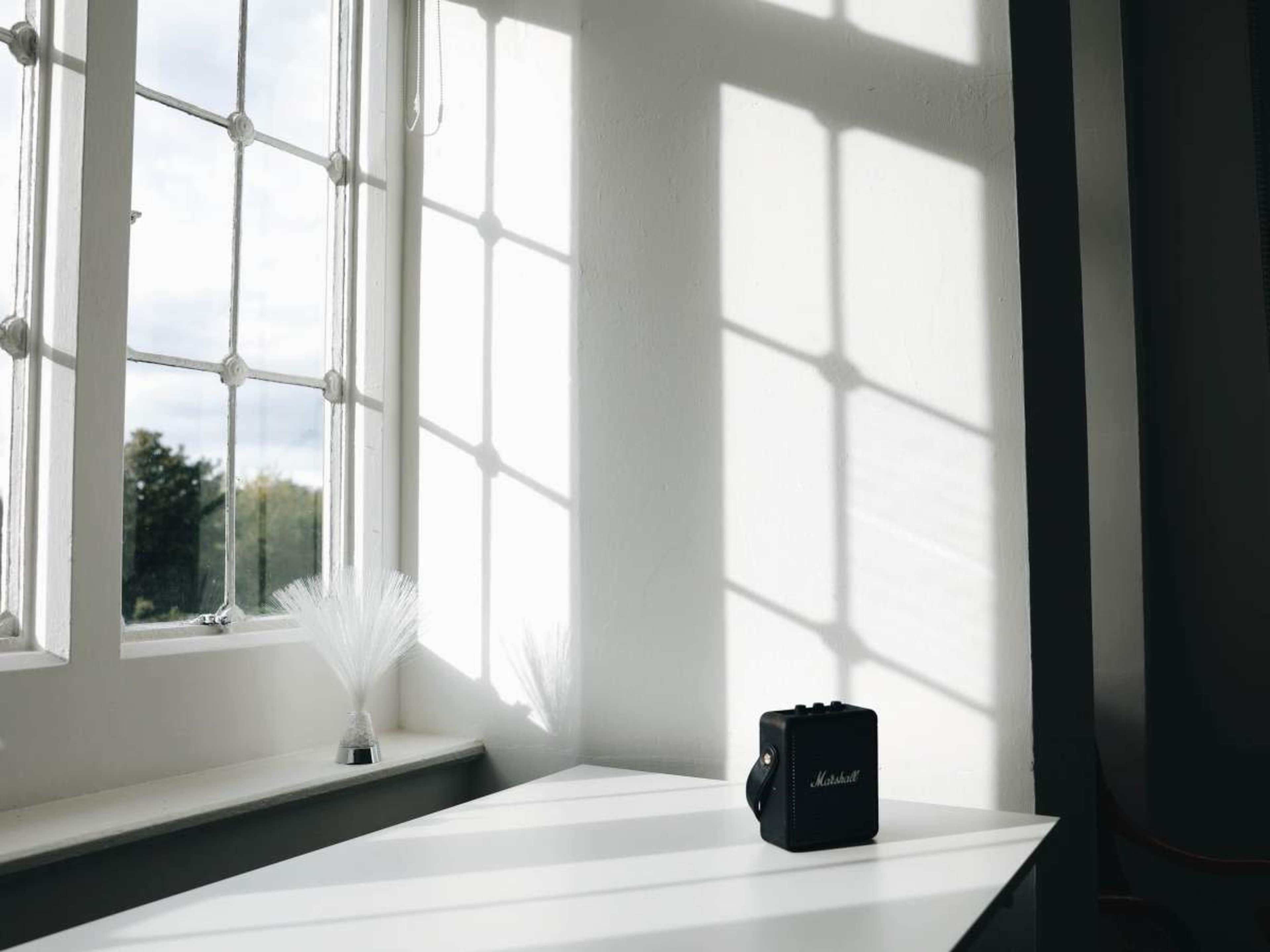 A black Marshall amplifier sits on a white table, illuminated by sunlight streaming through a window, casting shadows on the wall.