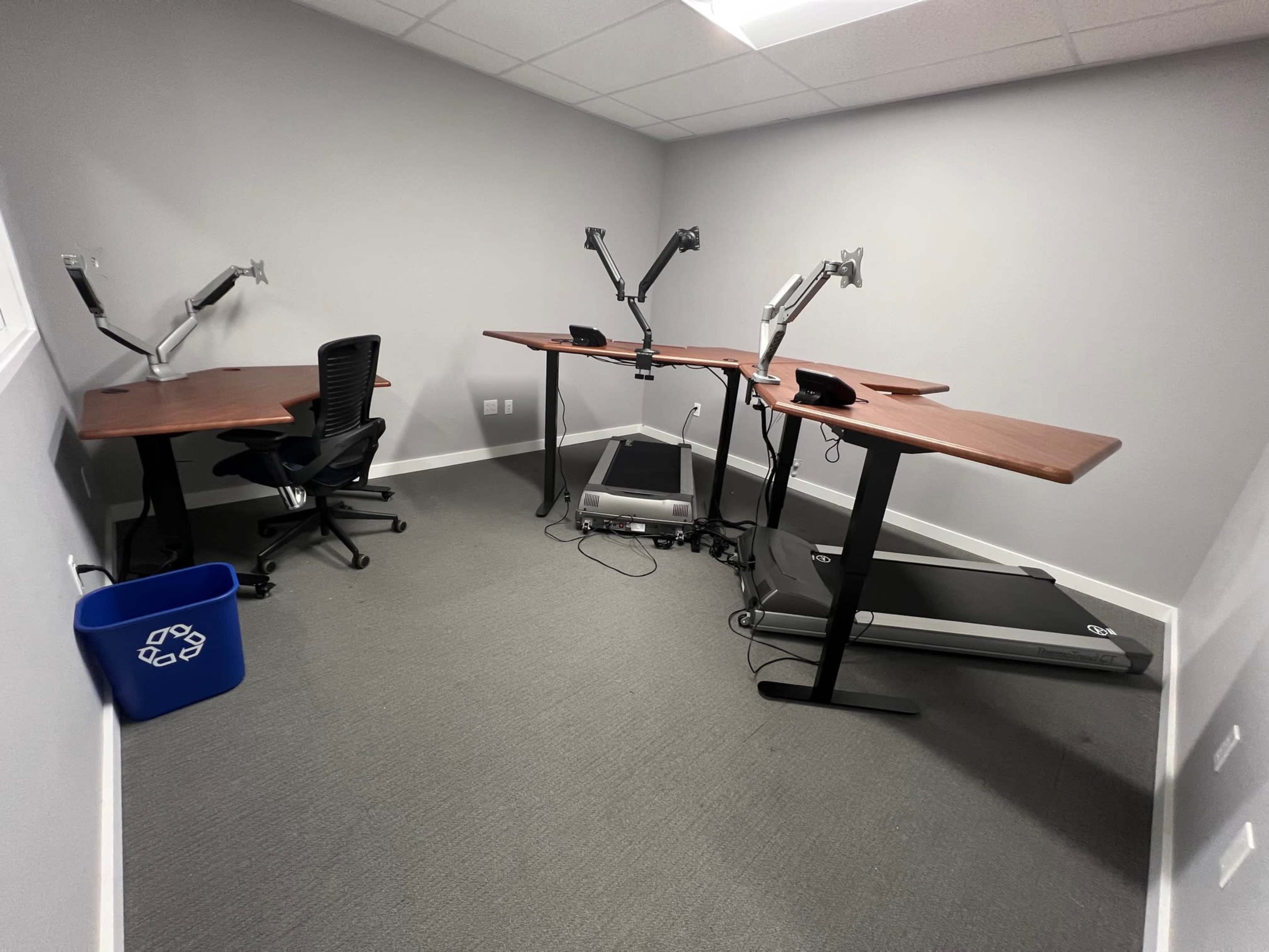 A small office space featuring two standing desks with adjustable arms, a black ergonomic chair, a treadmill, and a blue recycling bin.