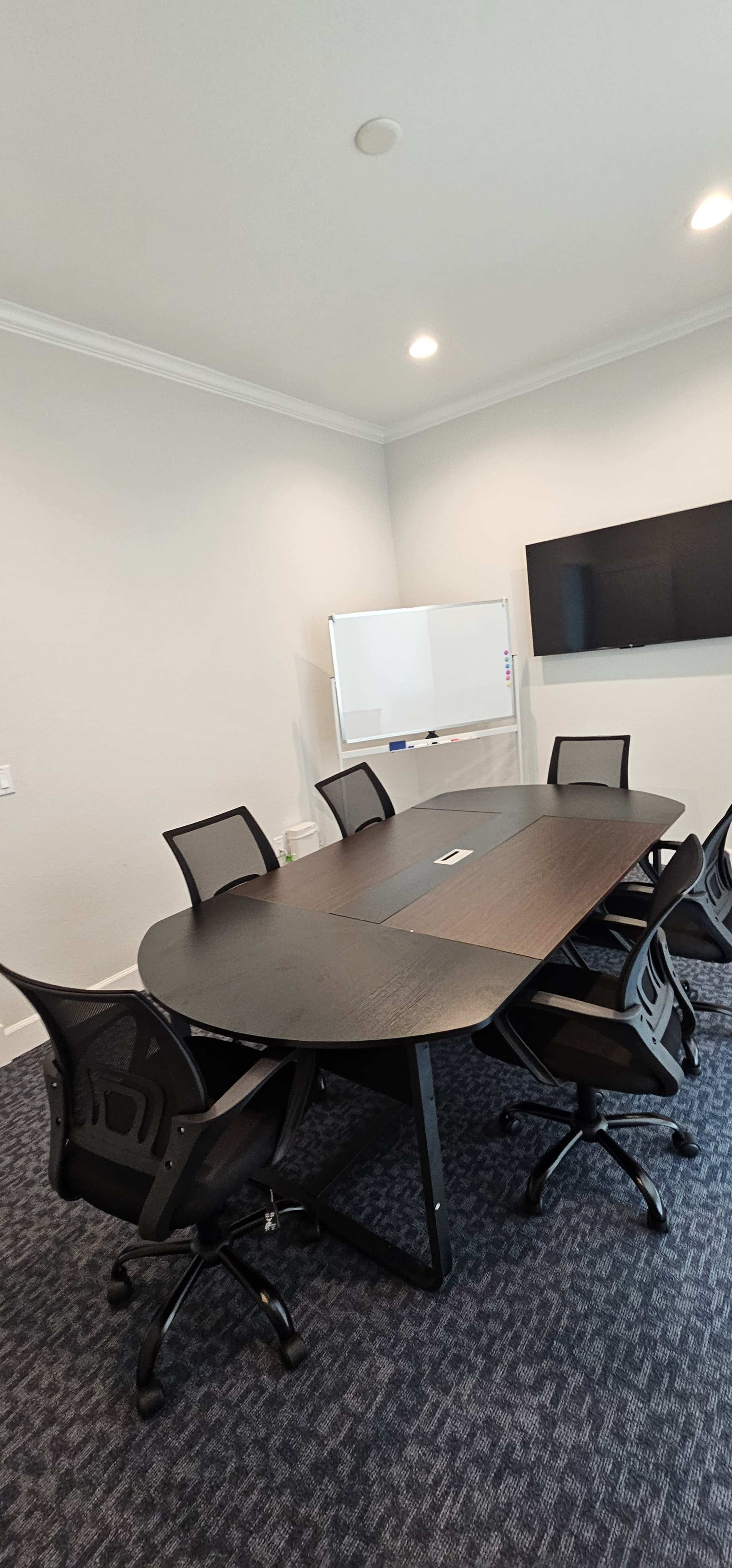 The image shows a modern conference room with an oval table surrounded by six ergonomic chairs, a whiteboard on one side, and a wall-mounted screen on the other.