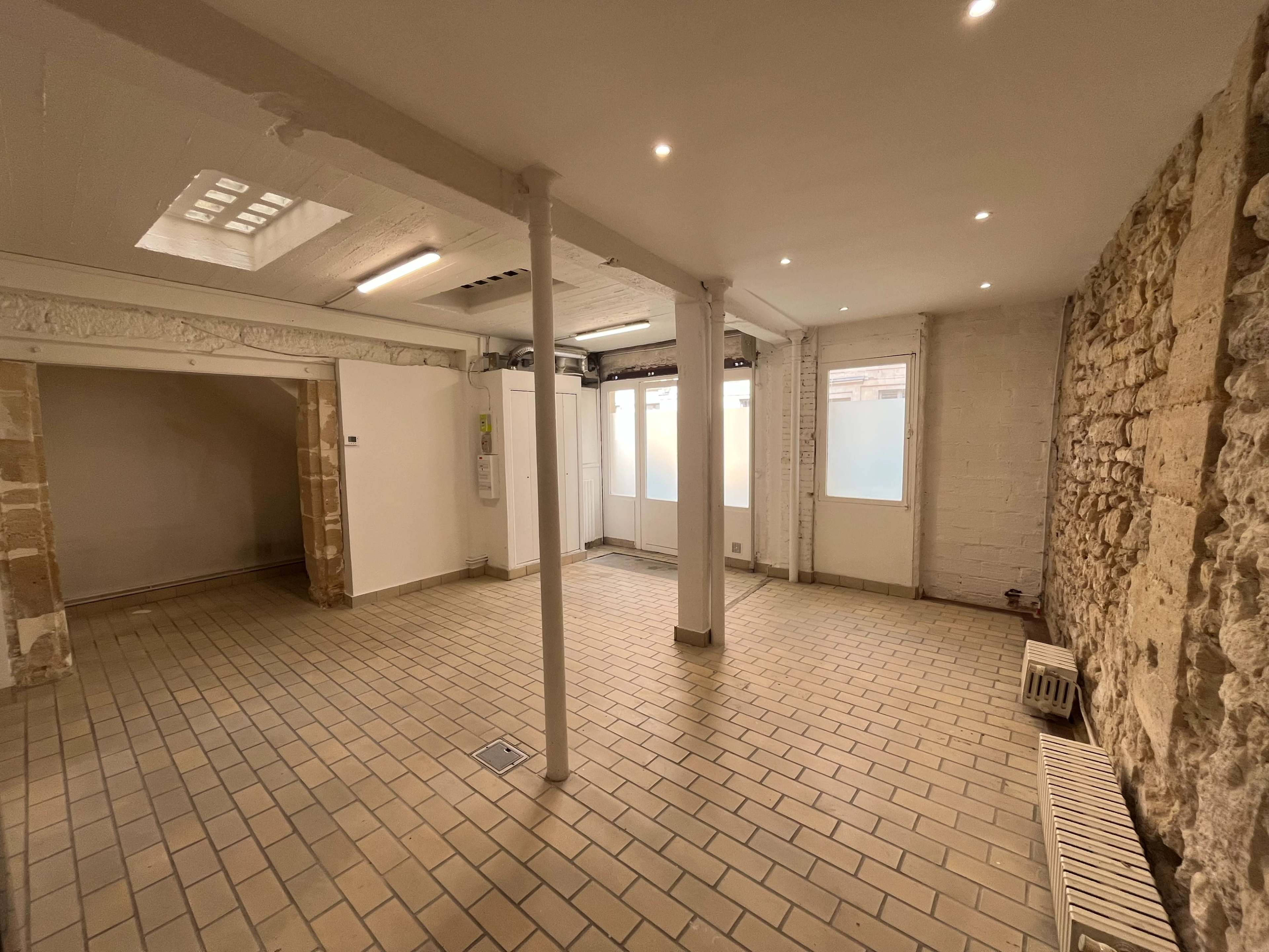 The image shows an empty, spacious interior room with stone and plaster walls, tiled flooring, and large windows.