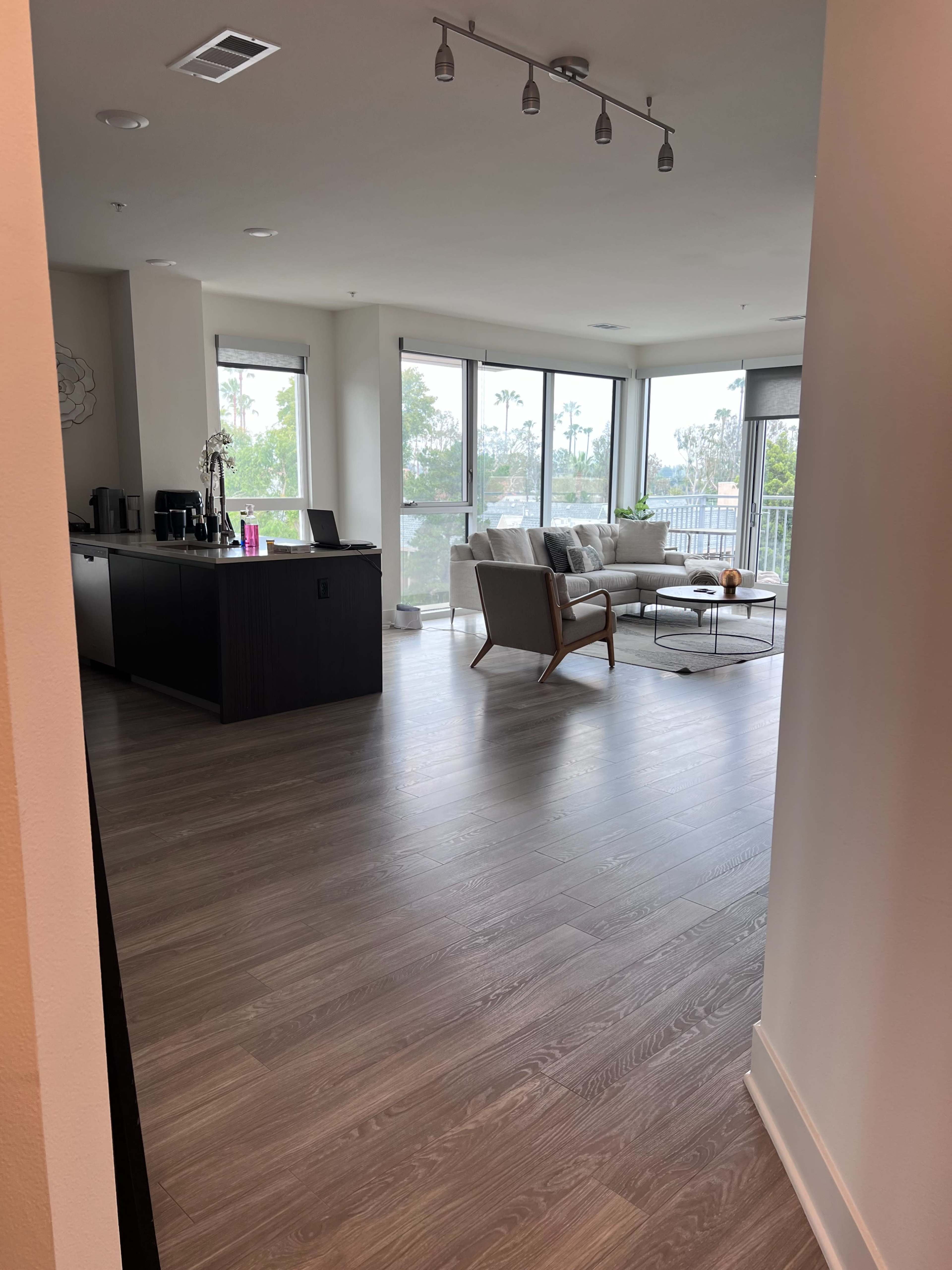 The image shows a modern open-concept living space with a view of large windows, a living area featuring a light-colored sofa, a coffee table, and an adjacent kitchen area.