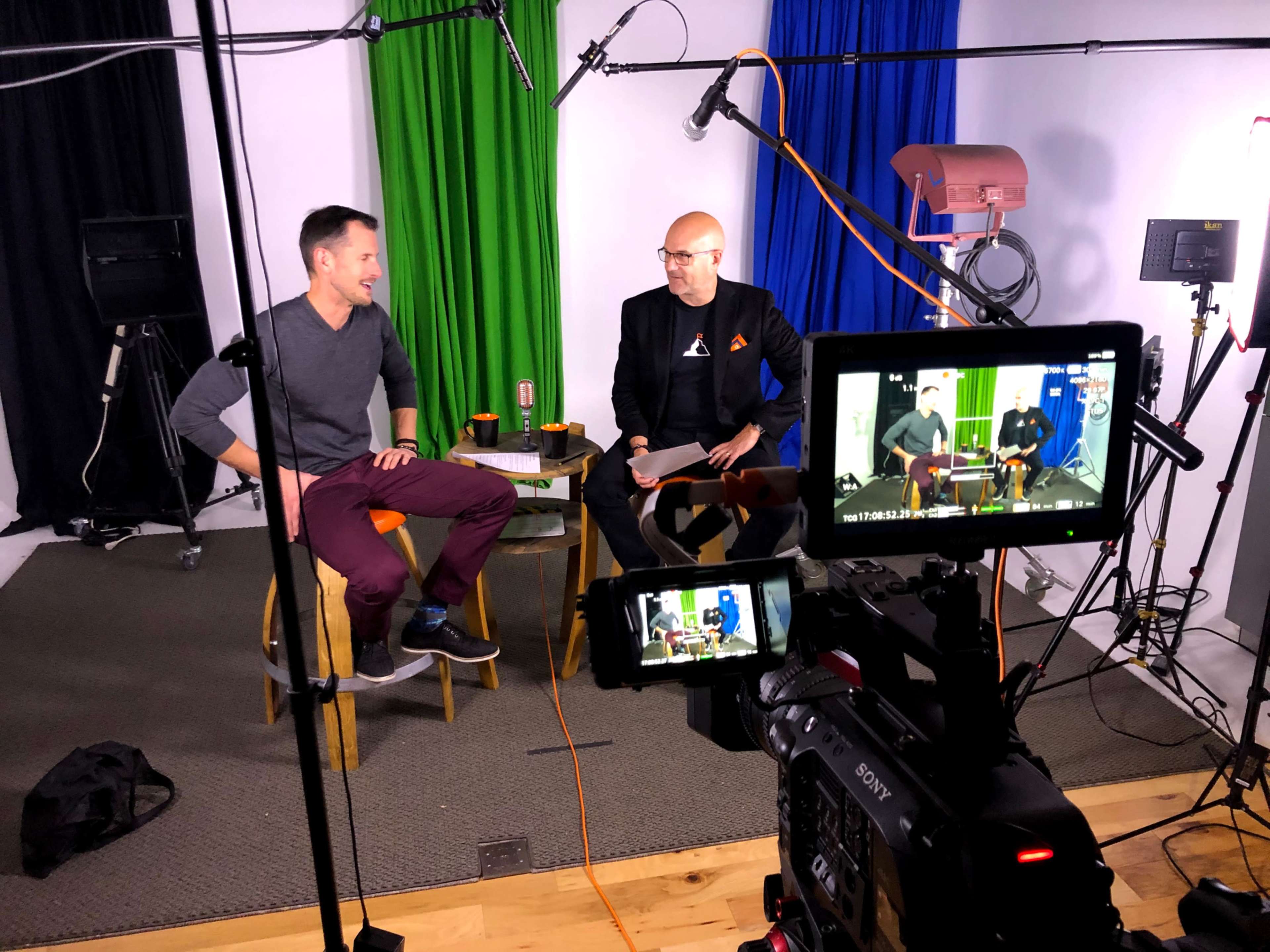 Two men are seated on stools in a studio setting equipped with multiple cameras and colorful backdrops.