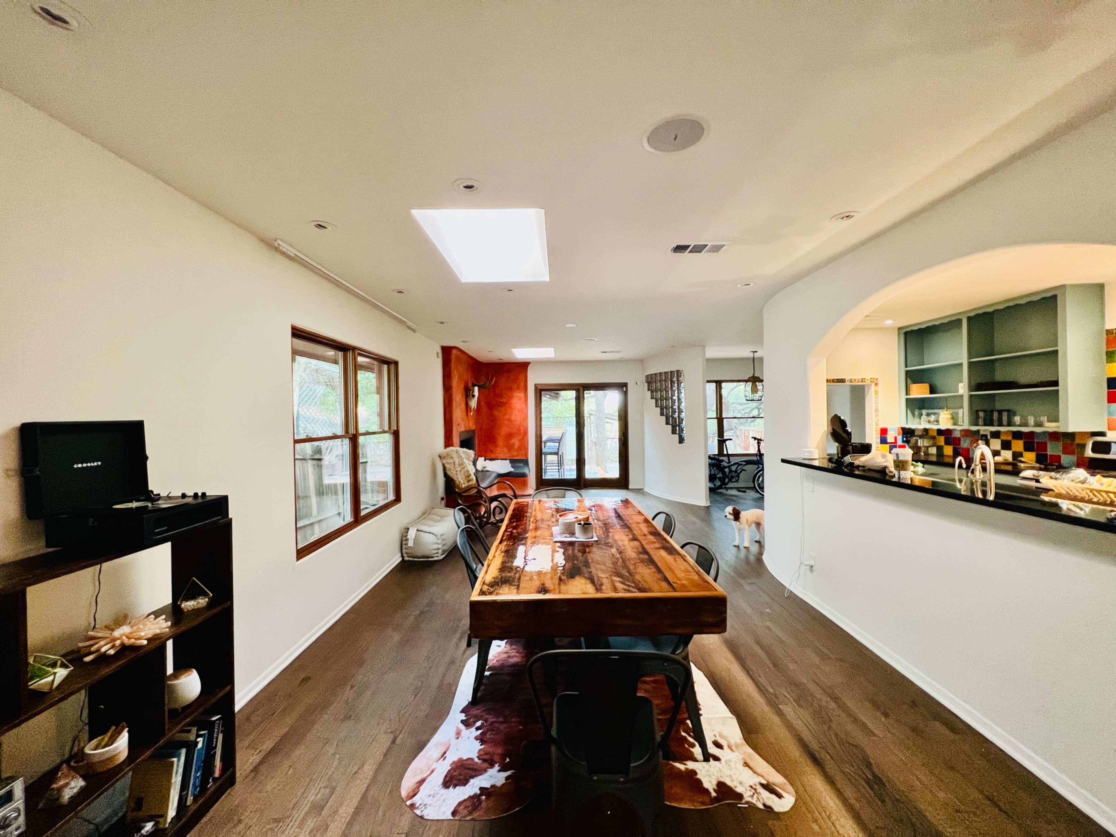 A spacious living area featuring a large wooden dining table, bright walls, and a kitchen visible in the background.