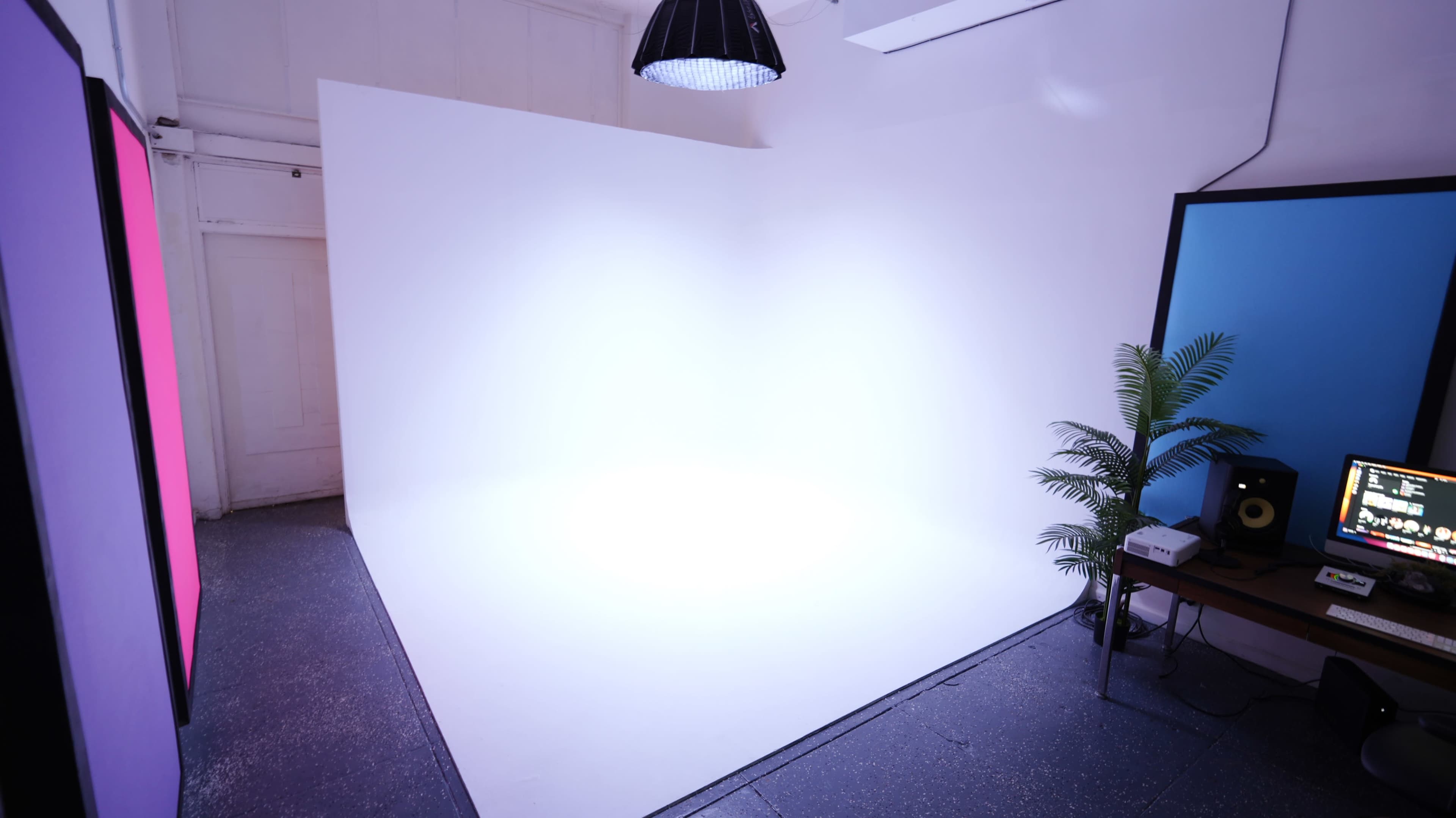The image shows a minimalistic interior space with a white backdrop, a pendant light, a small plant, and a table with equipment.