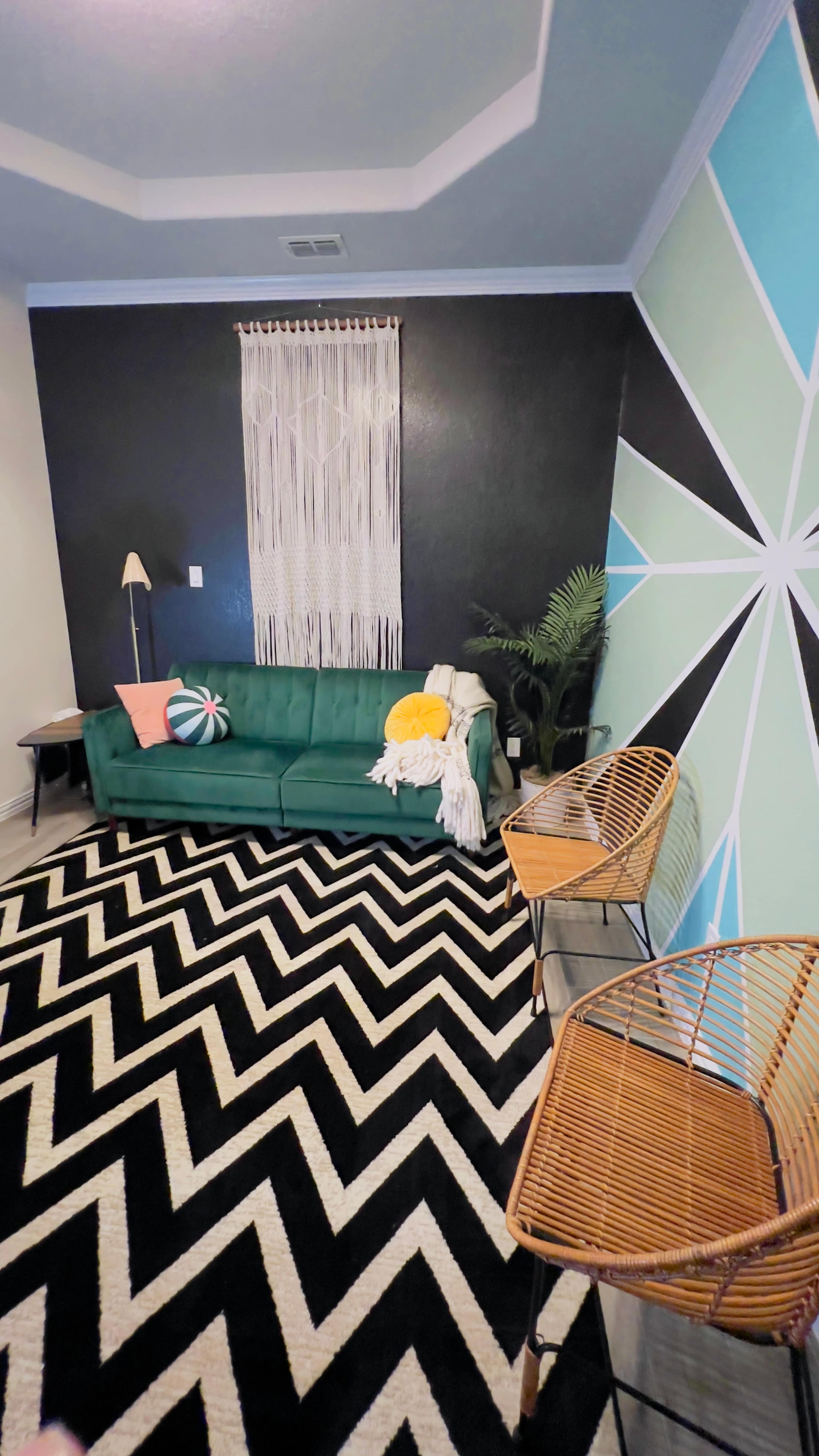 A modern living room featuring a green couch, two woven chairs, a patterned rug, and a colorful accent wall with geometric designs.