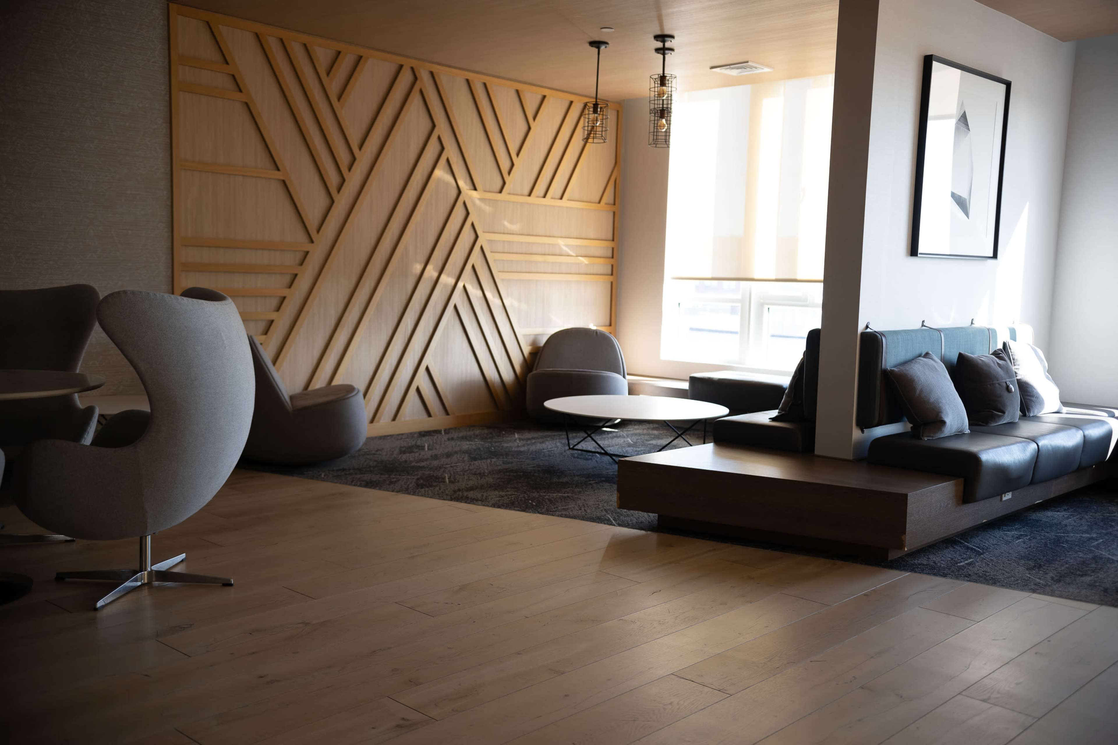 The image shows a modern lounge area featuring a wooden accent wall, a black leather sofa, a round coffee table, and several chairs arranged on a wooden floor.